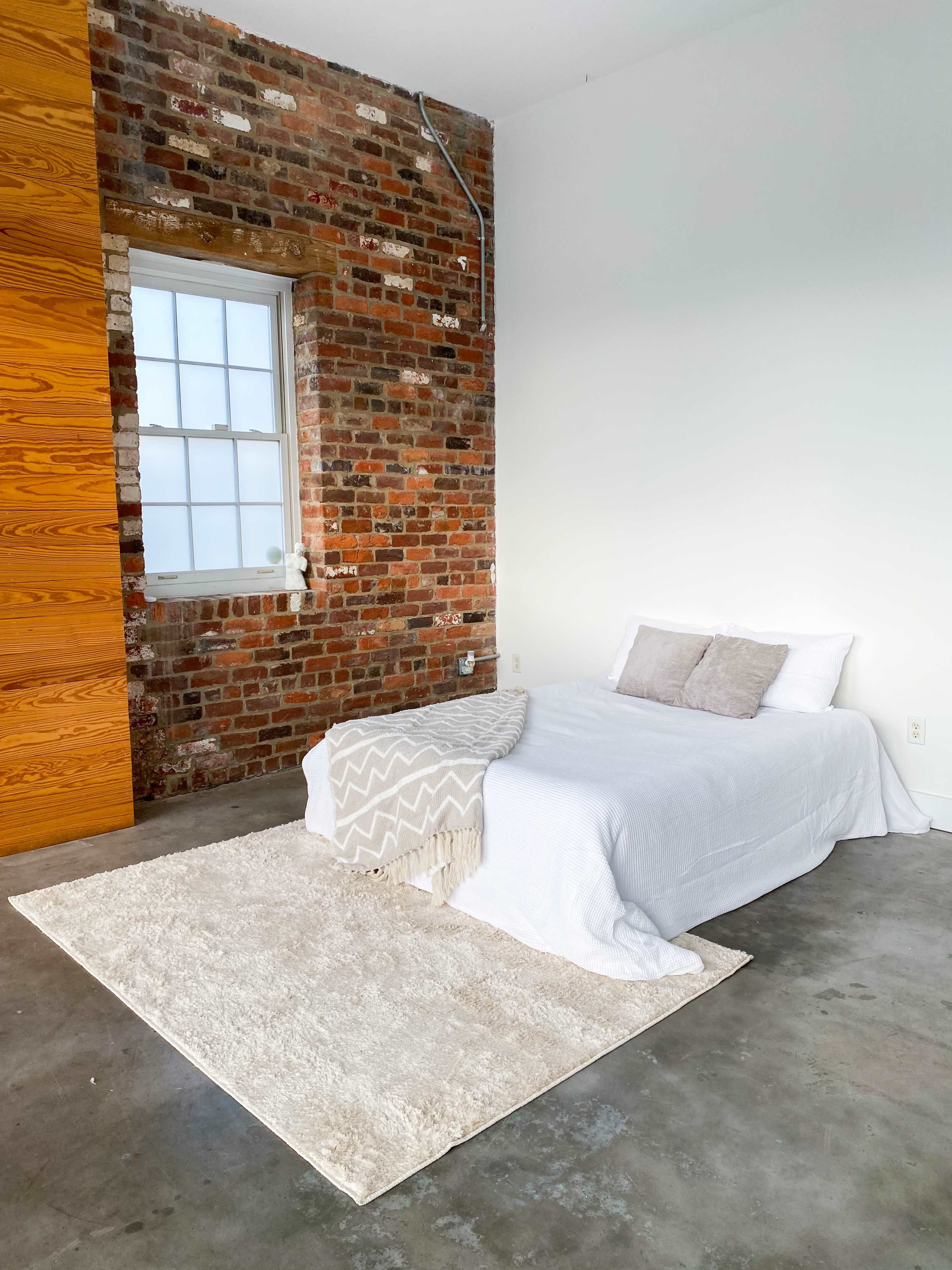The image shows a minimalist bedroom with a bed covered in white linens, a light-colored rug on a concrete floor, and a wall of exposed brick beside a window.