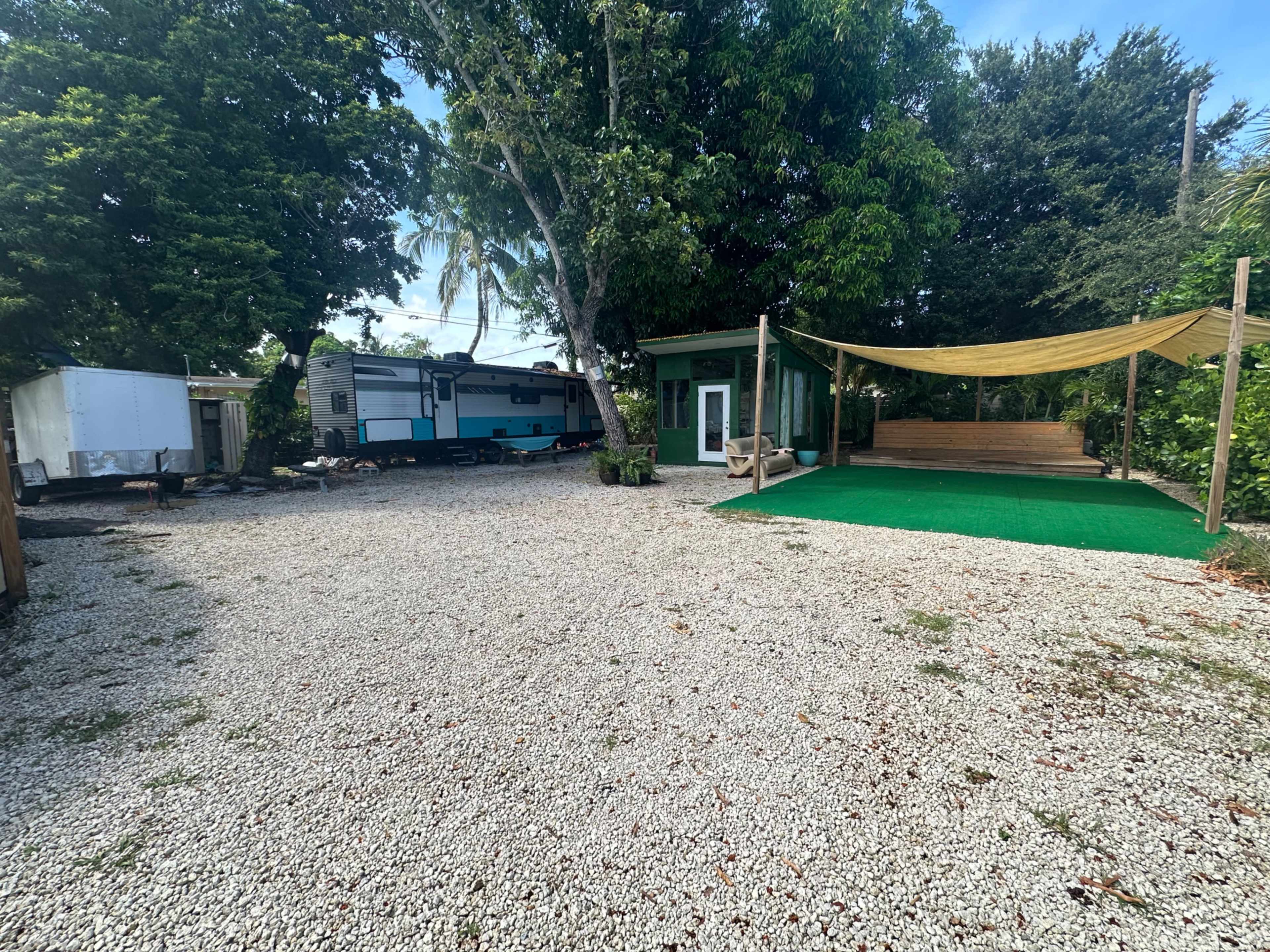 The image shows a gravel outdoor area featuring a green building, a parked trailer, and a covered outdoor seating space with artificial grass.