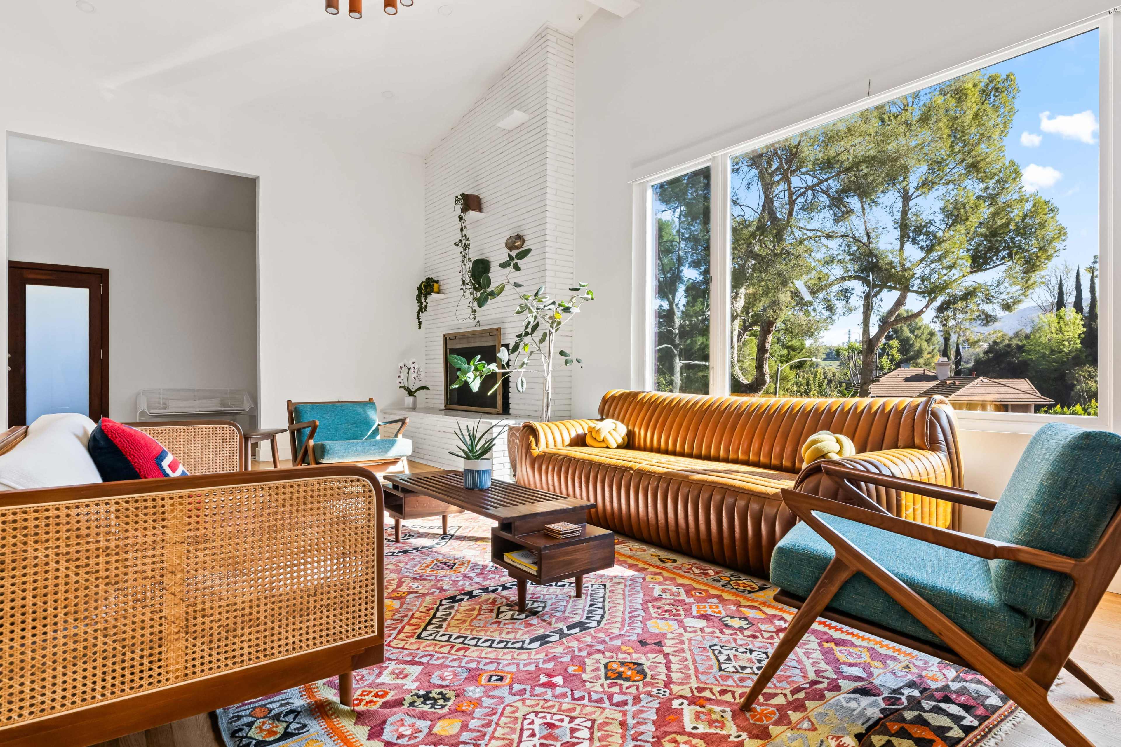 The brightly lit living room features a modern design with a leather couch, two teal armchairs, a wooden coffee table, and a colorful area rug, all overlooking a view of trees through large windows.