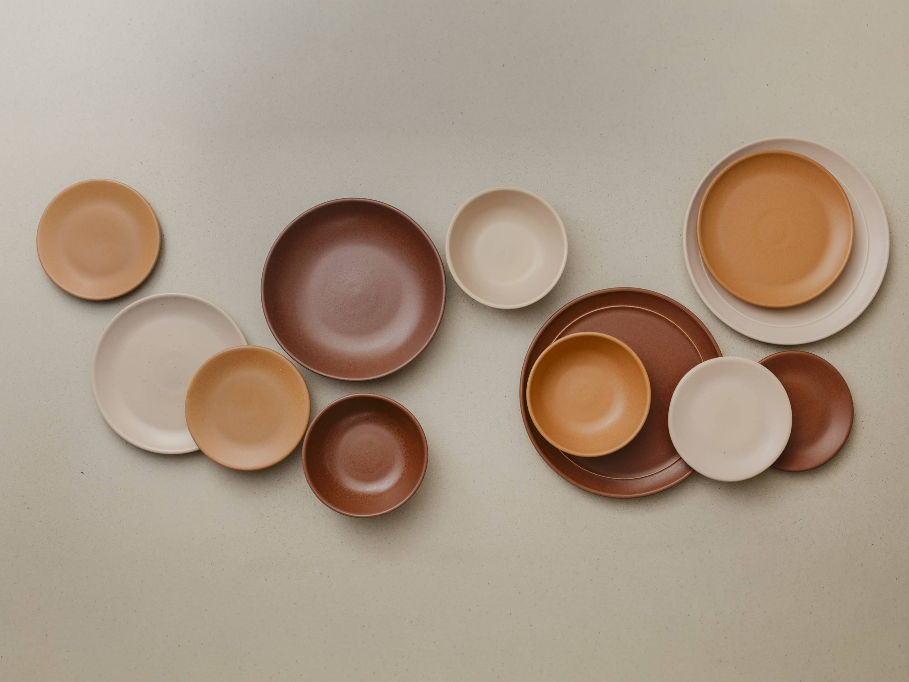 The image displays a collection of various ceramic plates and bowls in earthy tones arranged on a light background.