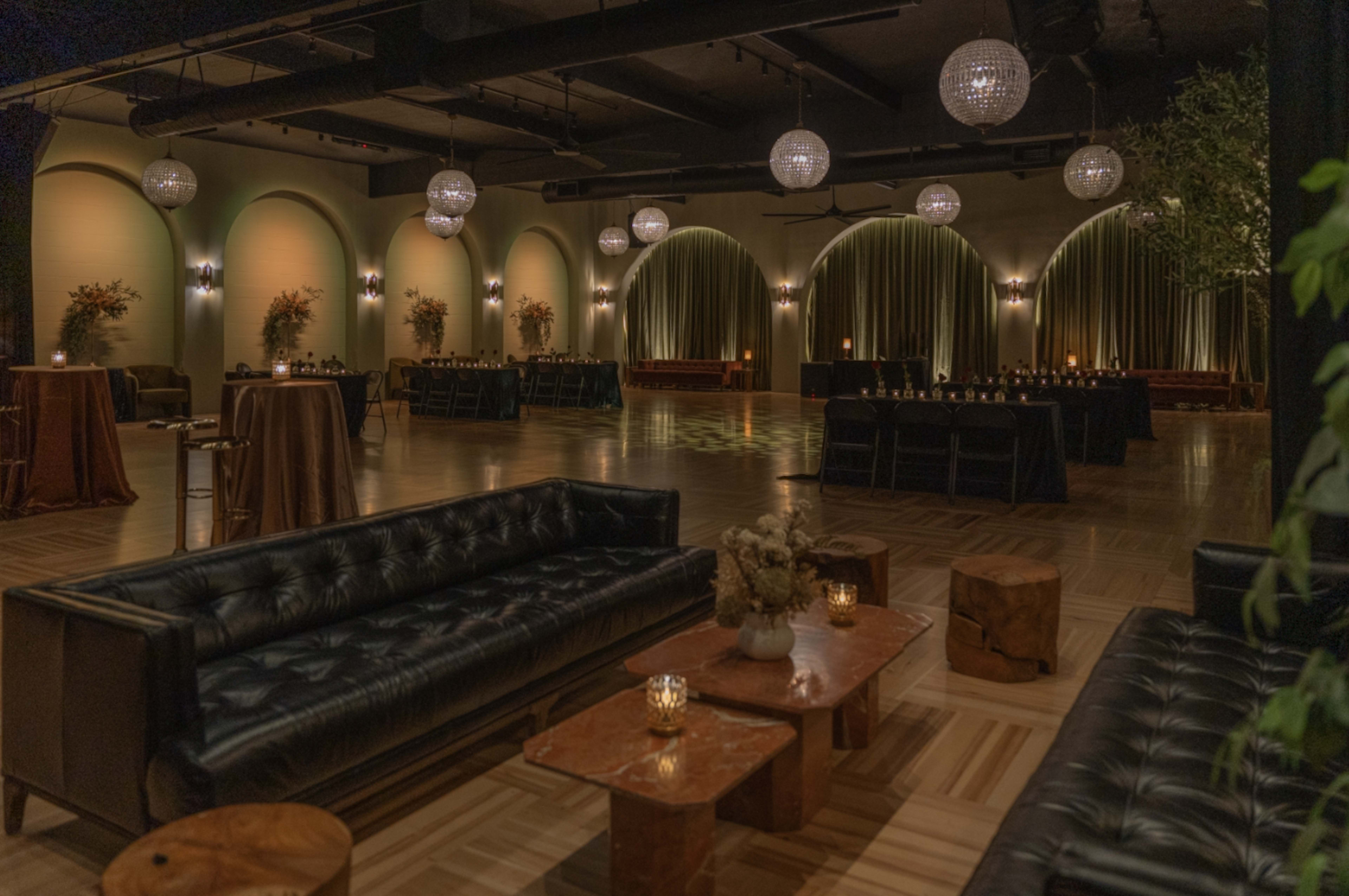 An elegantly arranged event space with dark sofas, wooden tables, and ambient lighting from chandeliers and candles.