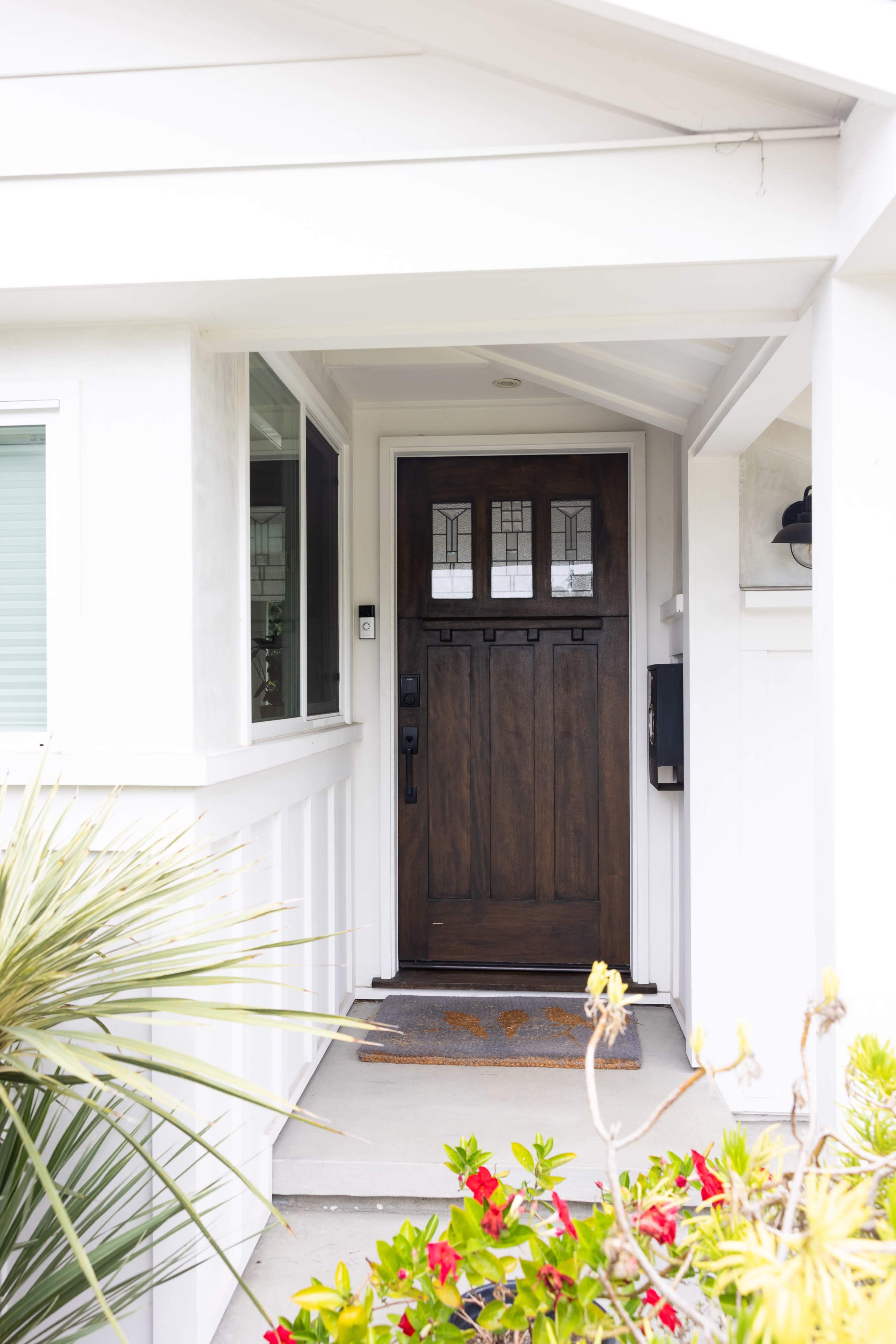 The image shows a residential entrance with a wooden front door framed by white walls and surrounded by plants and a welcome mat.