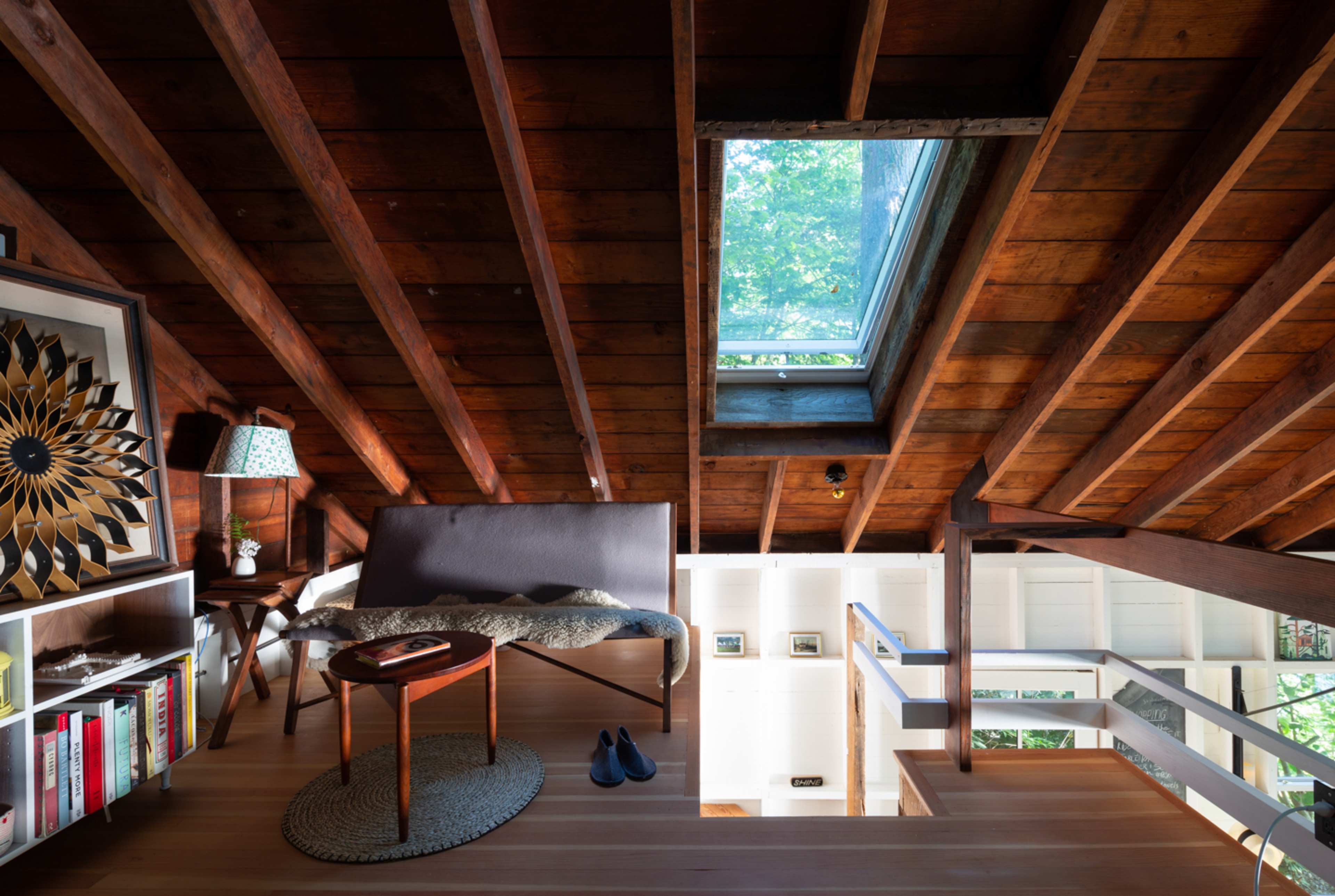 The image shows an open, rustic loft space with wooden beams, a skylight, a small sofa, a side table with a book, and a few decorative items.