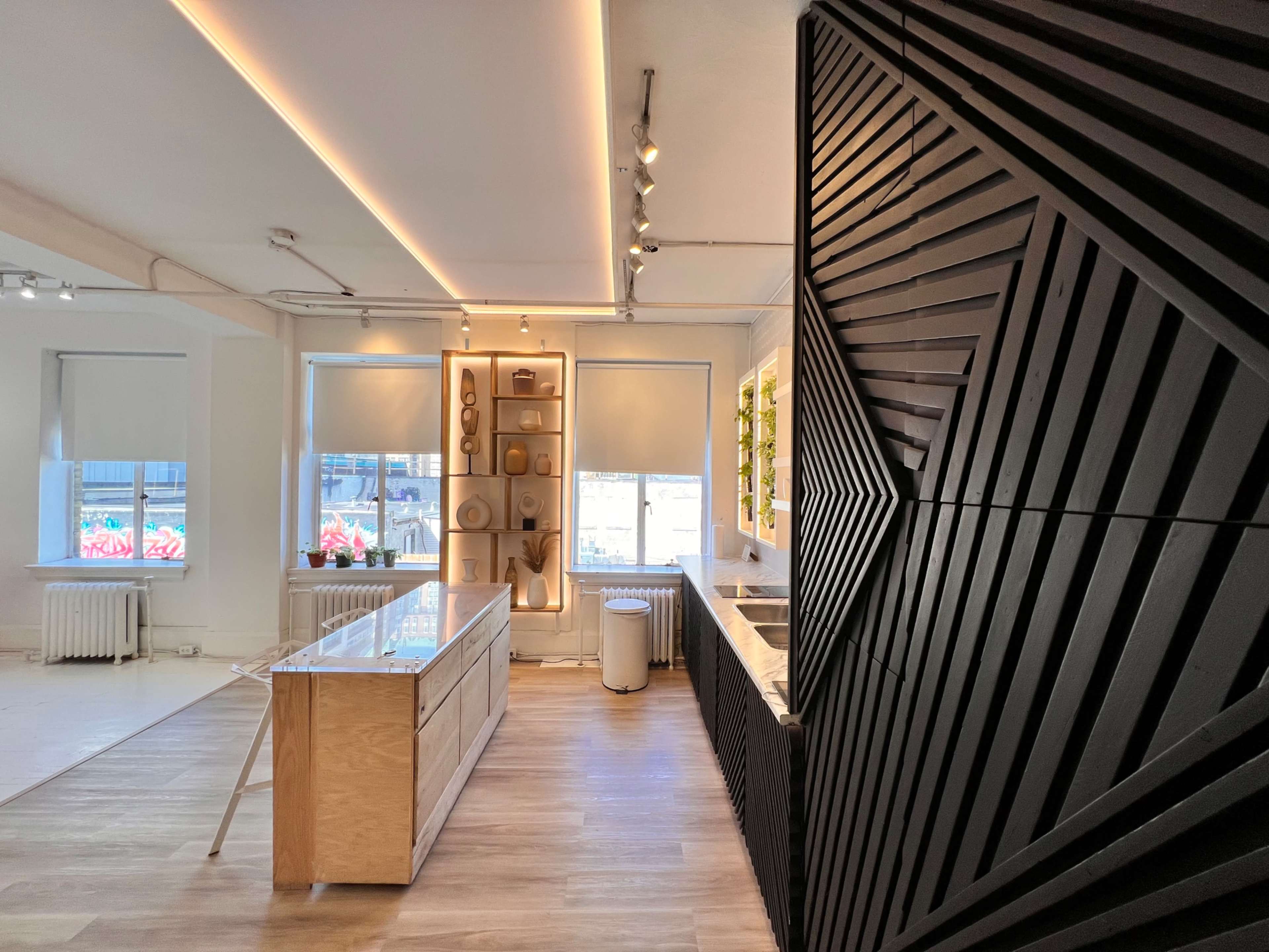 A modern kitchen featuring a wooden cabinet unit, black geometric wall paneling, and large windows allowing natural light.