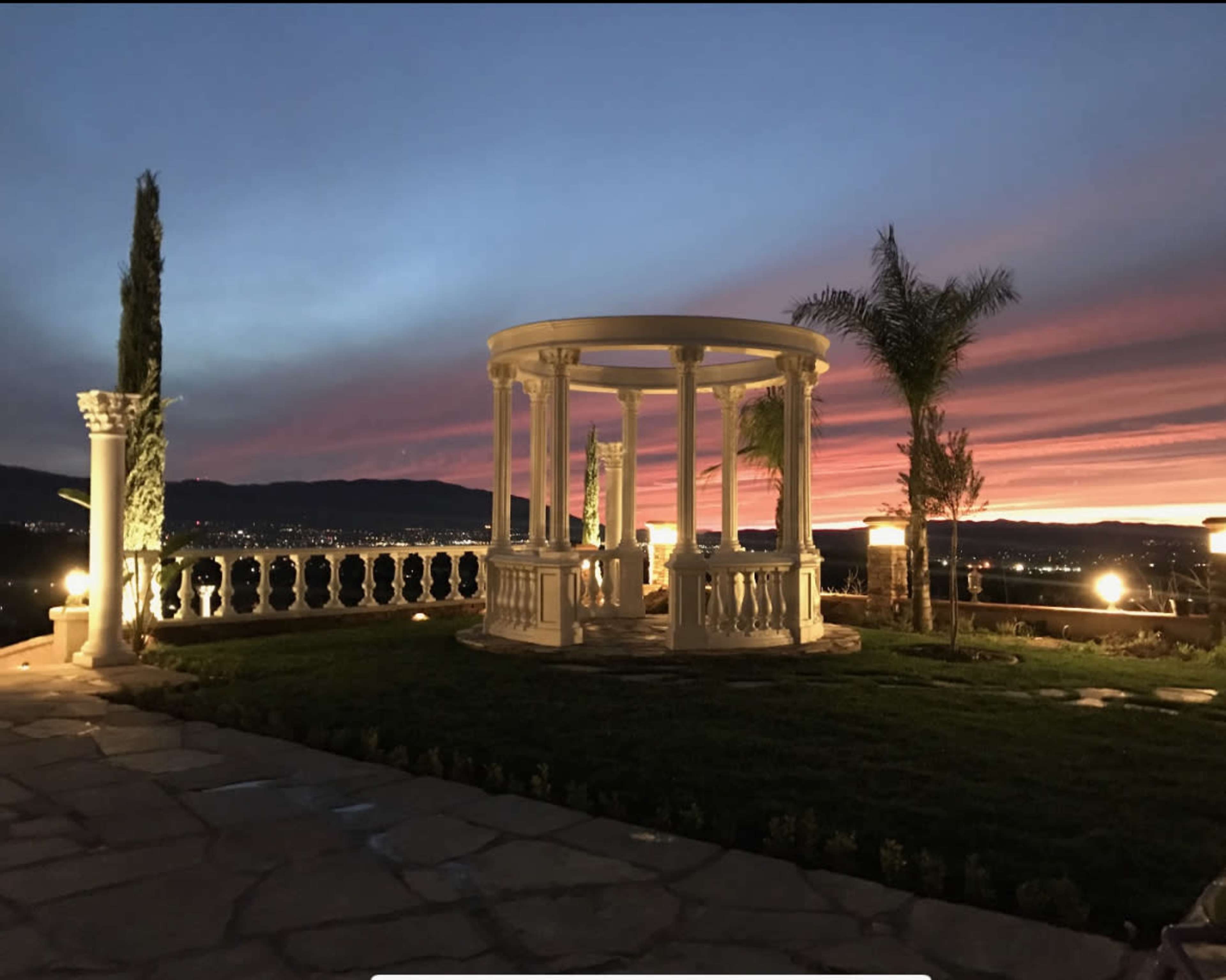A white gazebo is seated on a stone patio, surrounded by manicured lawns and palm trees, with a vibrant sunset casting colorful hues in the sky.