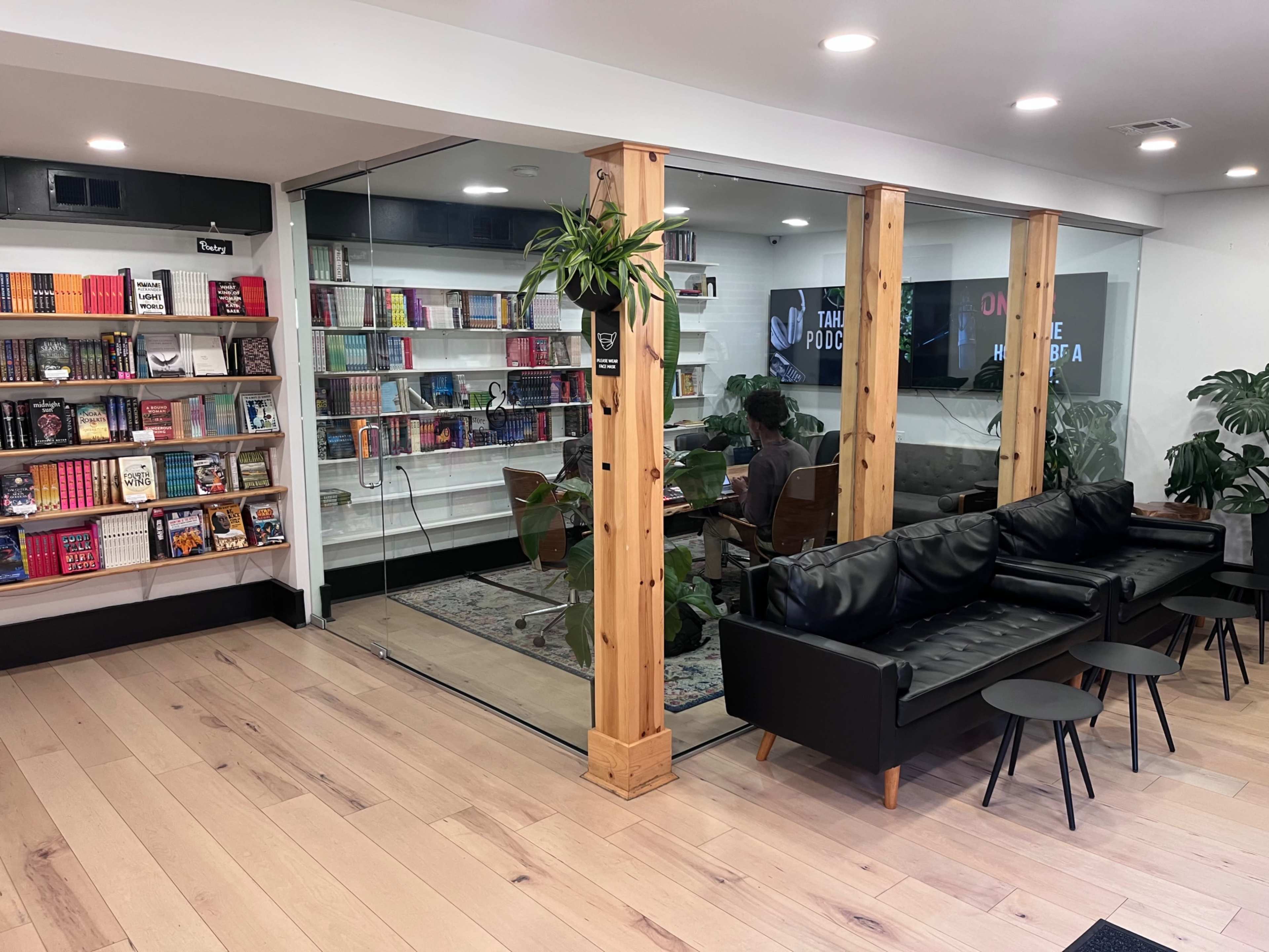 The image shows a cozy reading space with bookshelves on one side and a glass-walled meeting area on the other, featuring a sofa and small tables.
