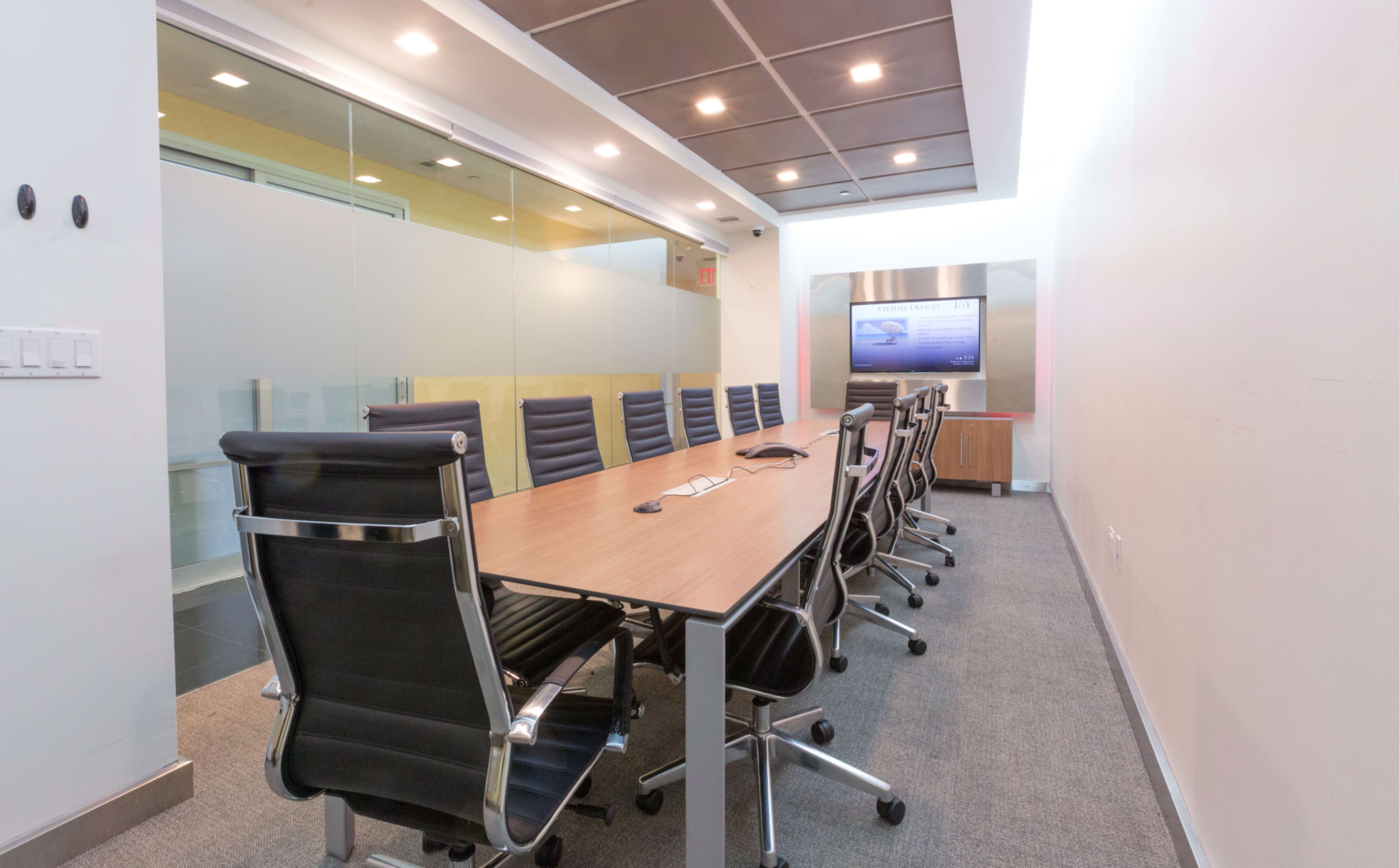 A modern conference room features a long wooden table surrounded by black office chairs, with a large screen and glass walls.