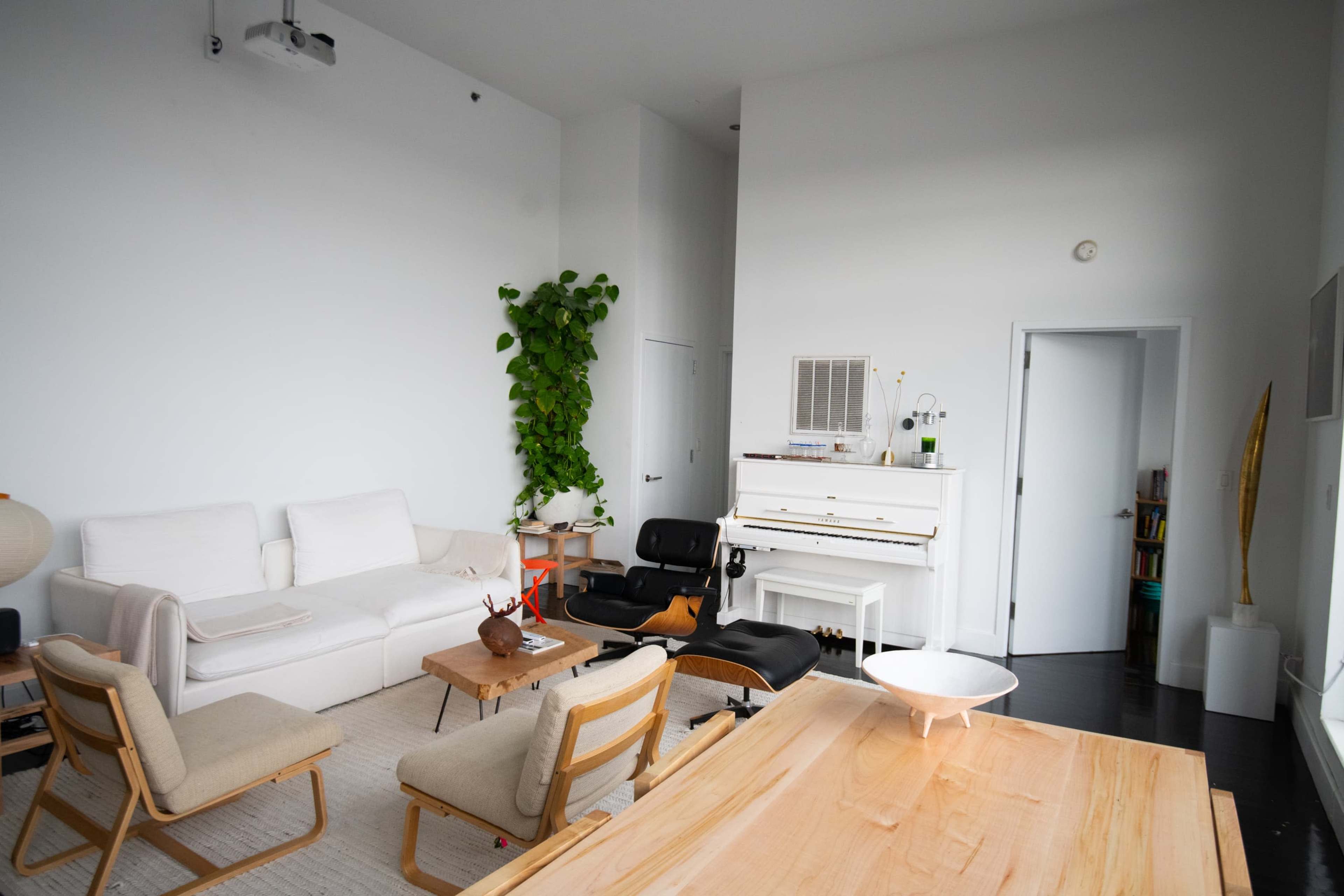 A modern, minimalist living room featuring a white sofa, wooden table, potted green plant, and a piano against a white wall.