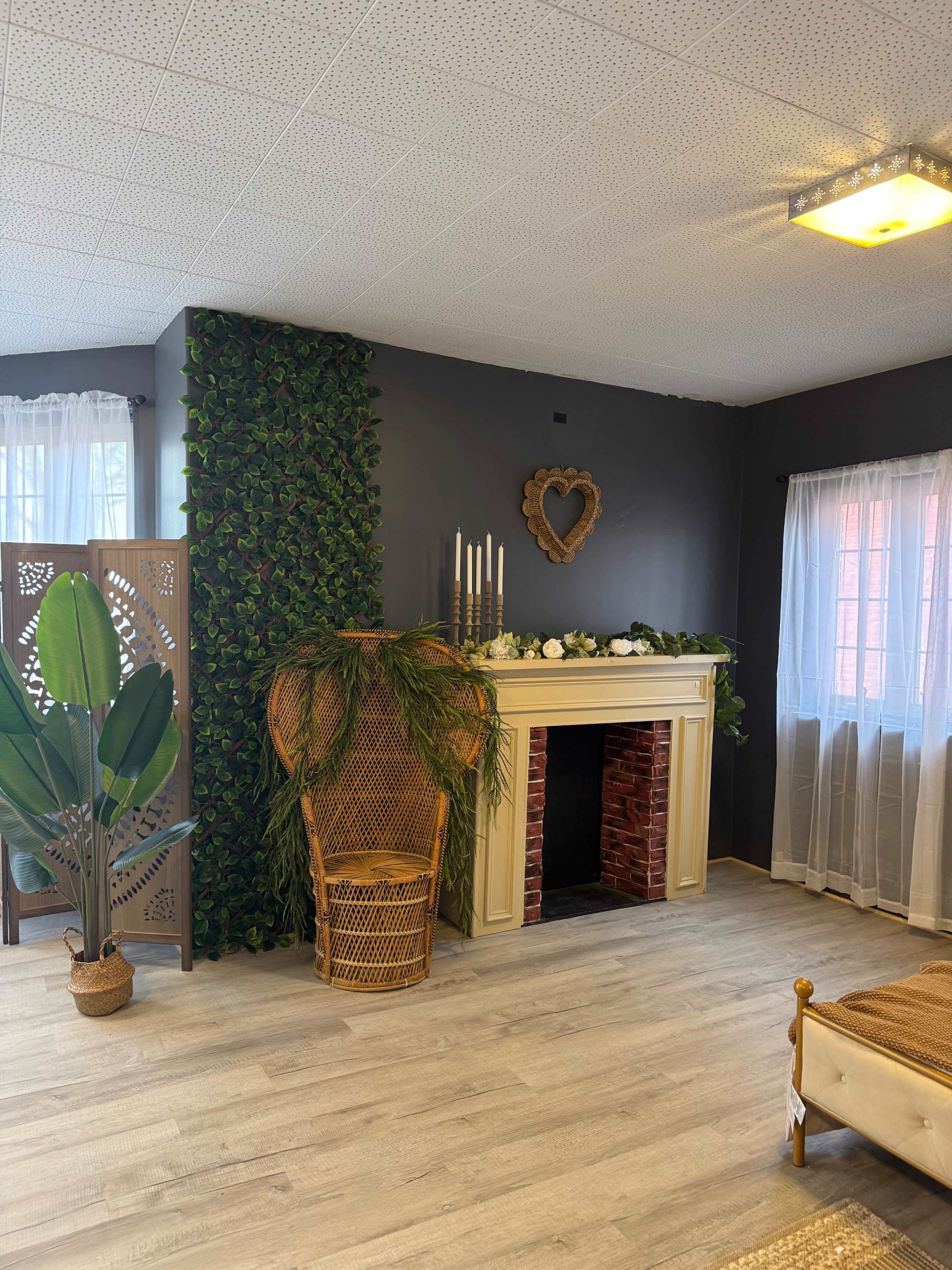 The image shows a softly lit room with a decorative faux fireplace, a woven chair, green plants on the walls, and sheer white curtains.