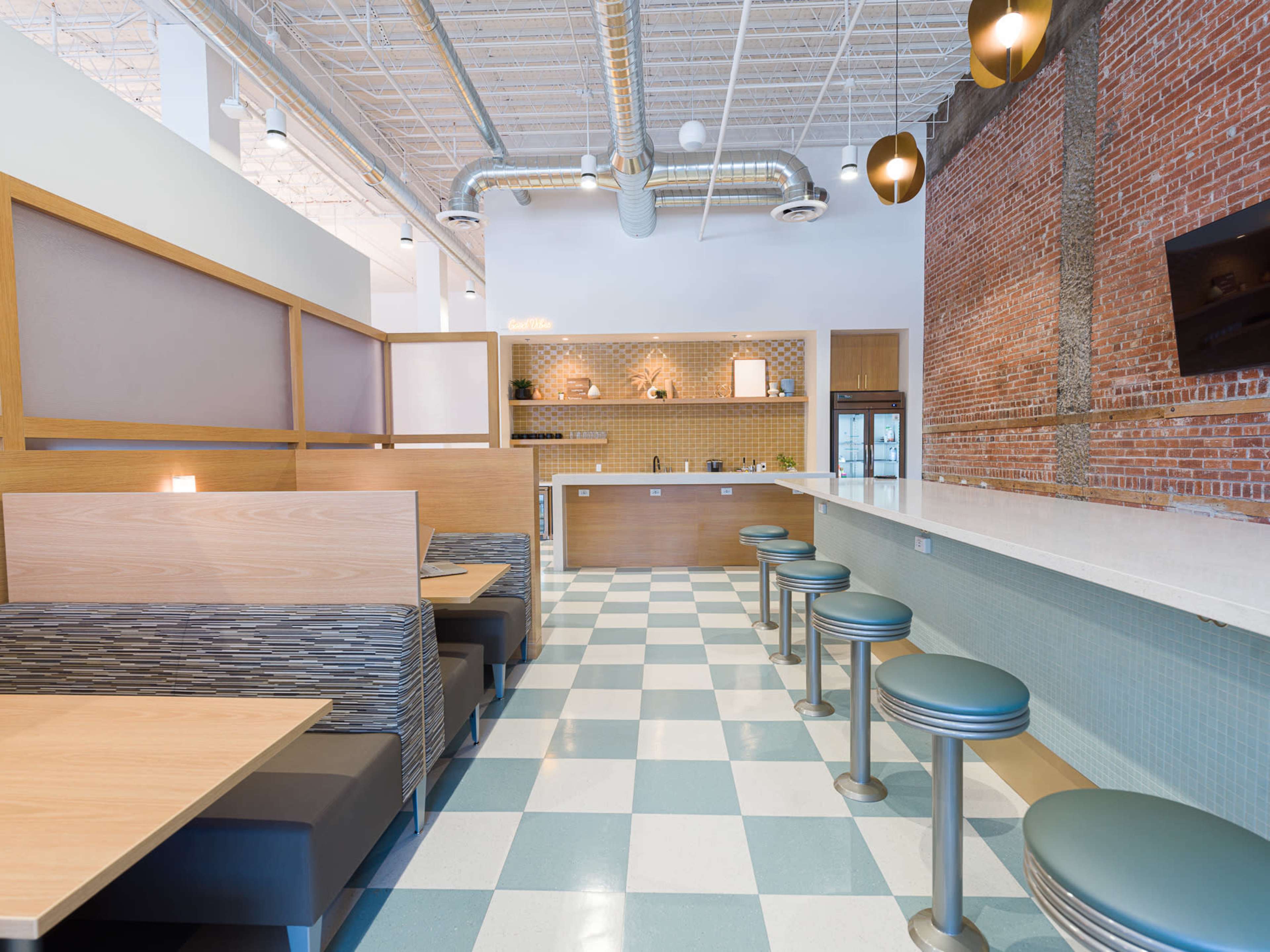The image shows a modern café interior with booth seating and a countertop bar area featuring round stools, brick and tiled walls, and open kitchen space.