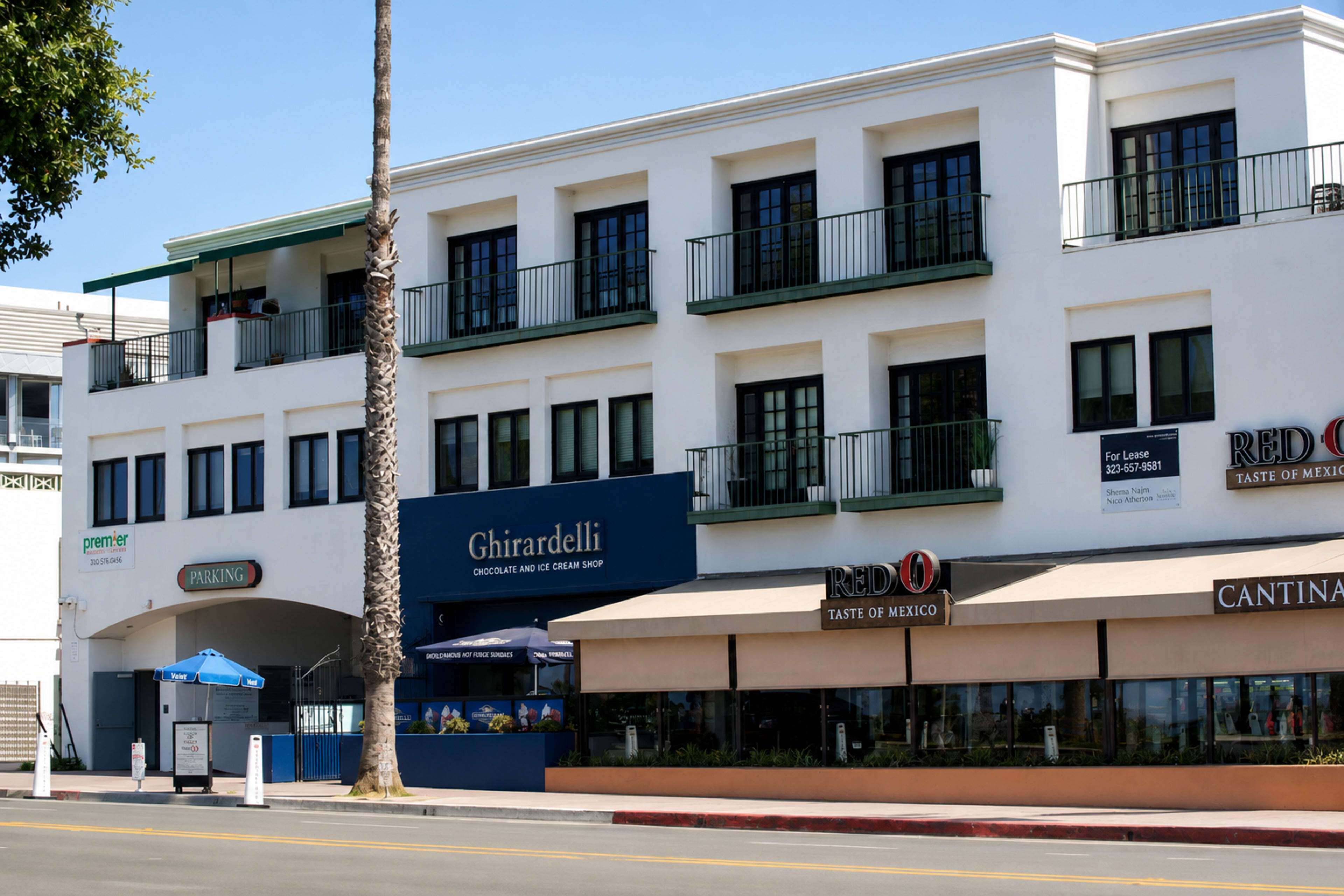 The image shows a two-story building with businesses on the ground floor, including a Ghirardelli chocolate shop and a restaurant, situated along a street with palm trees.