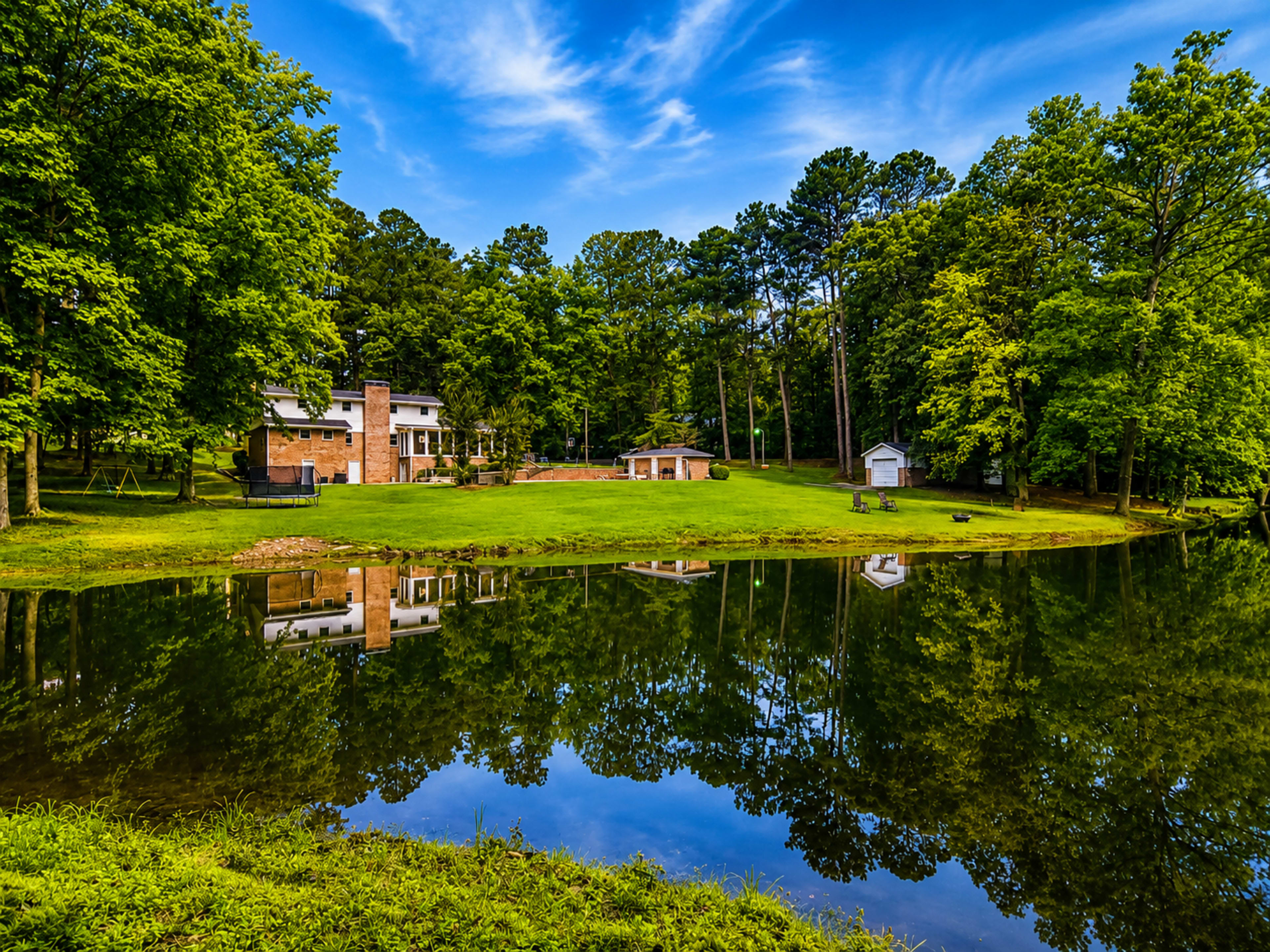 A picturesque house with a brick facade is surrounded by trees and reflected in a calm pond, with a swing set and a small shed visible in the background.