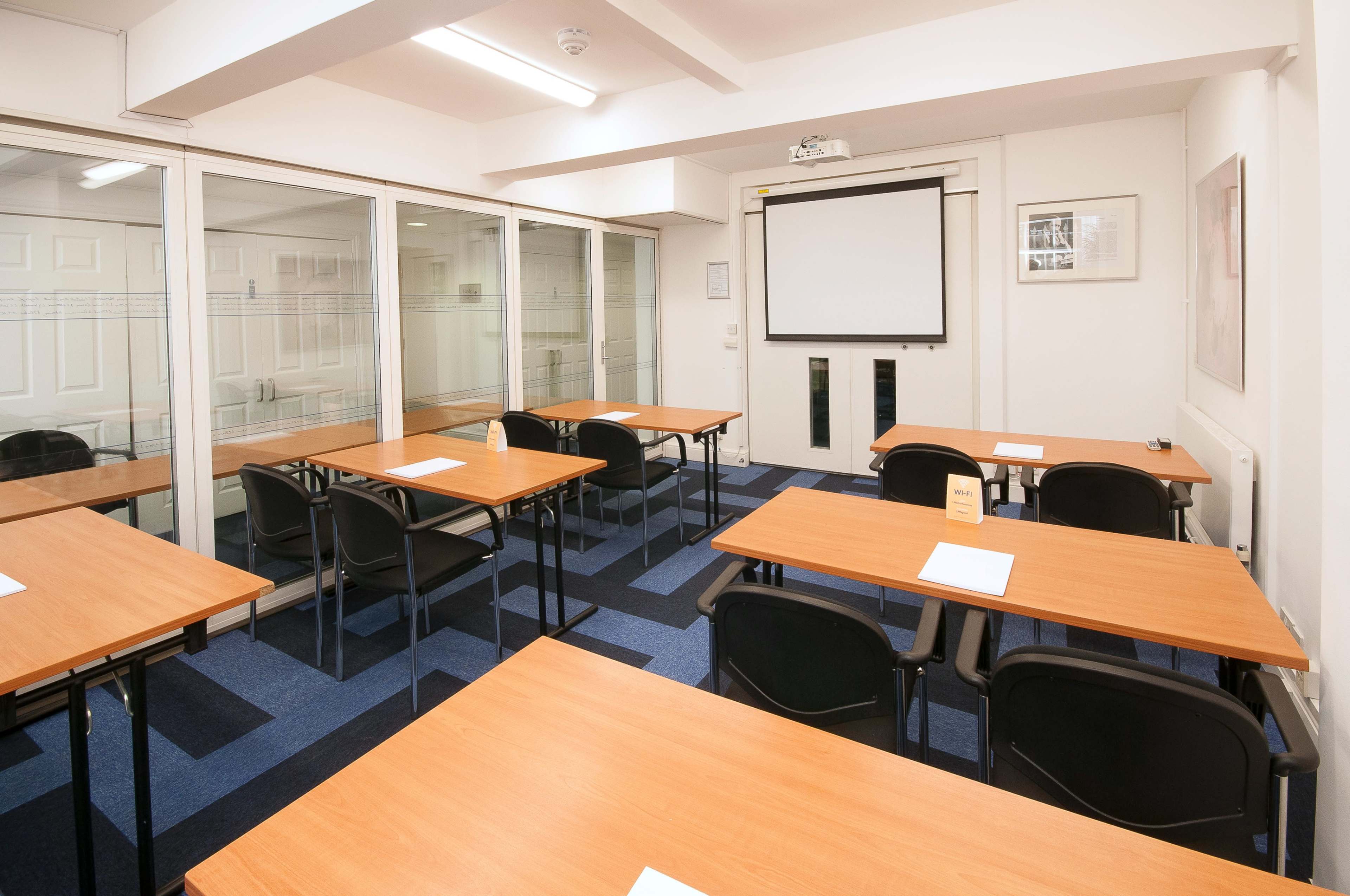 A conference room features several wooden tables and chairs arranged with a projector screen at the front.