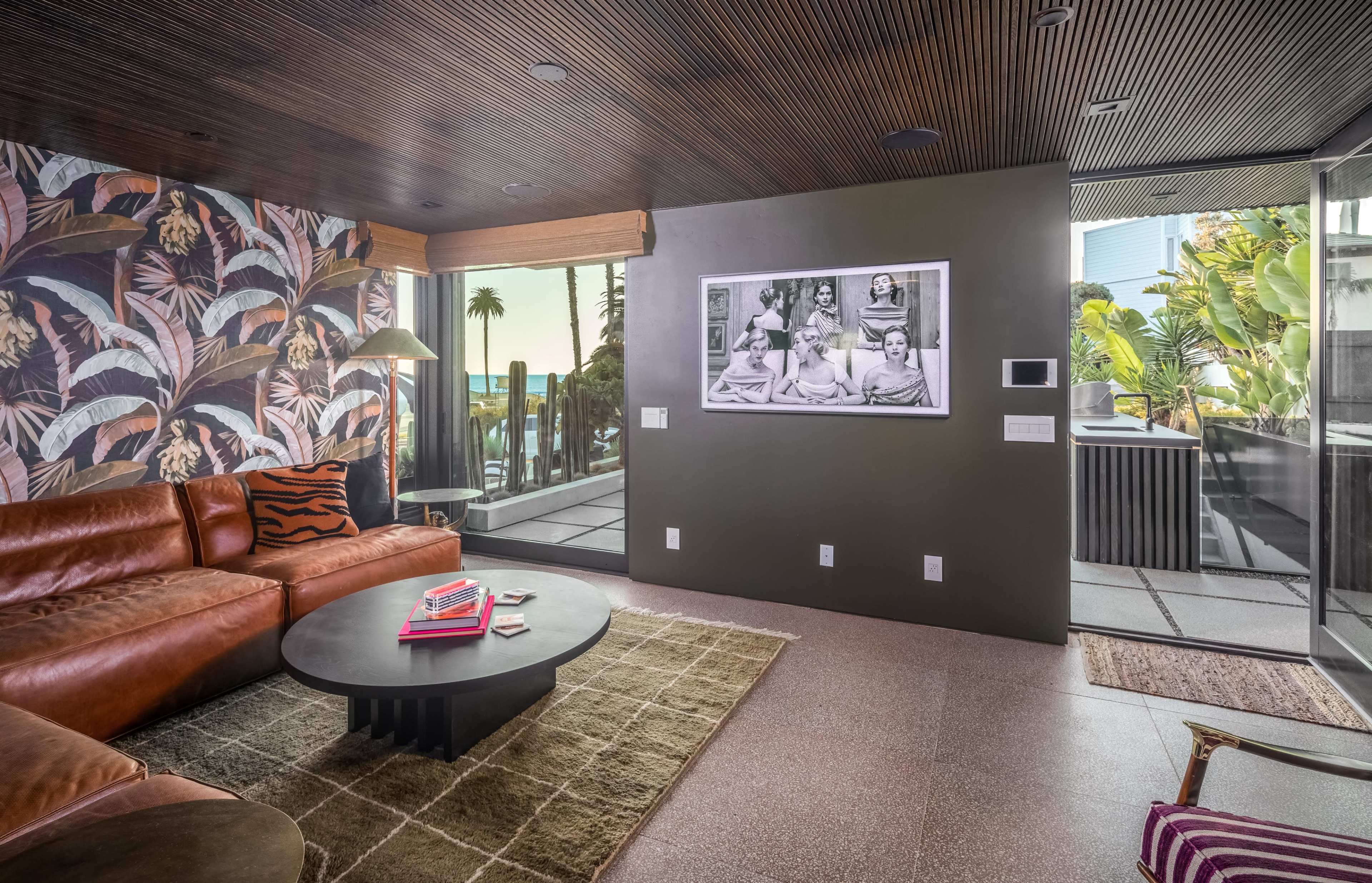 A modern living room featuring a leather sofa, a round coffee table, and a large wall with a black and white photograph, opening to an outdoor space with palm trees.