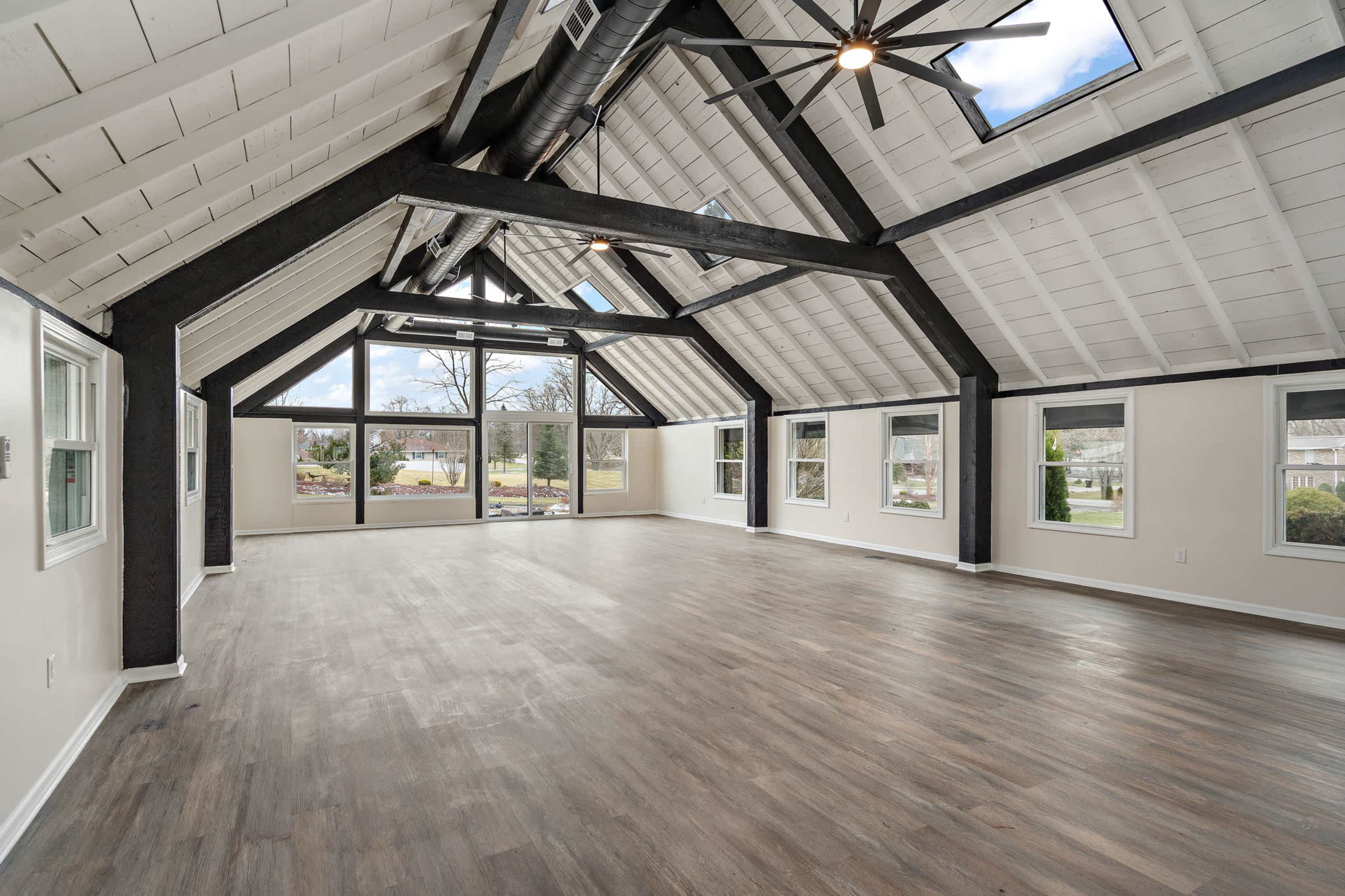 The image depicts a spacious, well-lit interior room with high vaulted ceilings, large windows, and a wooden floor.
