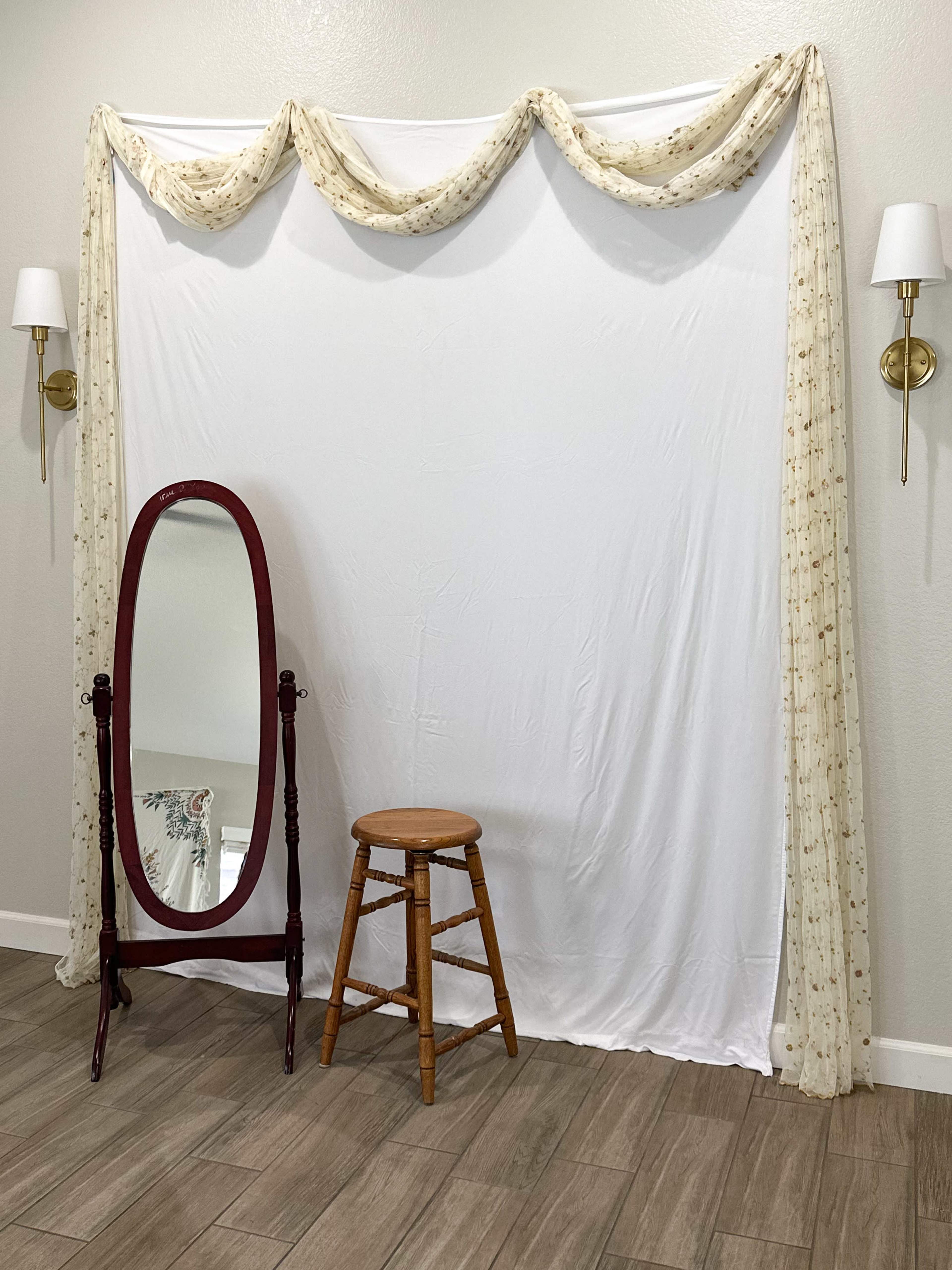 A white backdrop is draped with fabric at the top, accompanied by a wooden stool and a full-length mirror on the floor.
