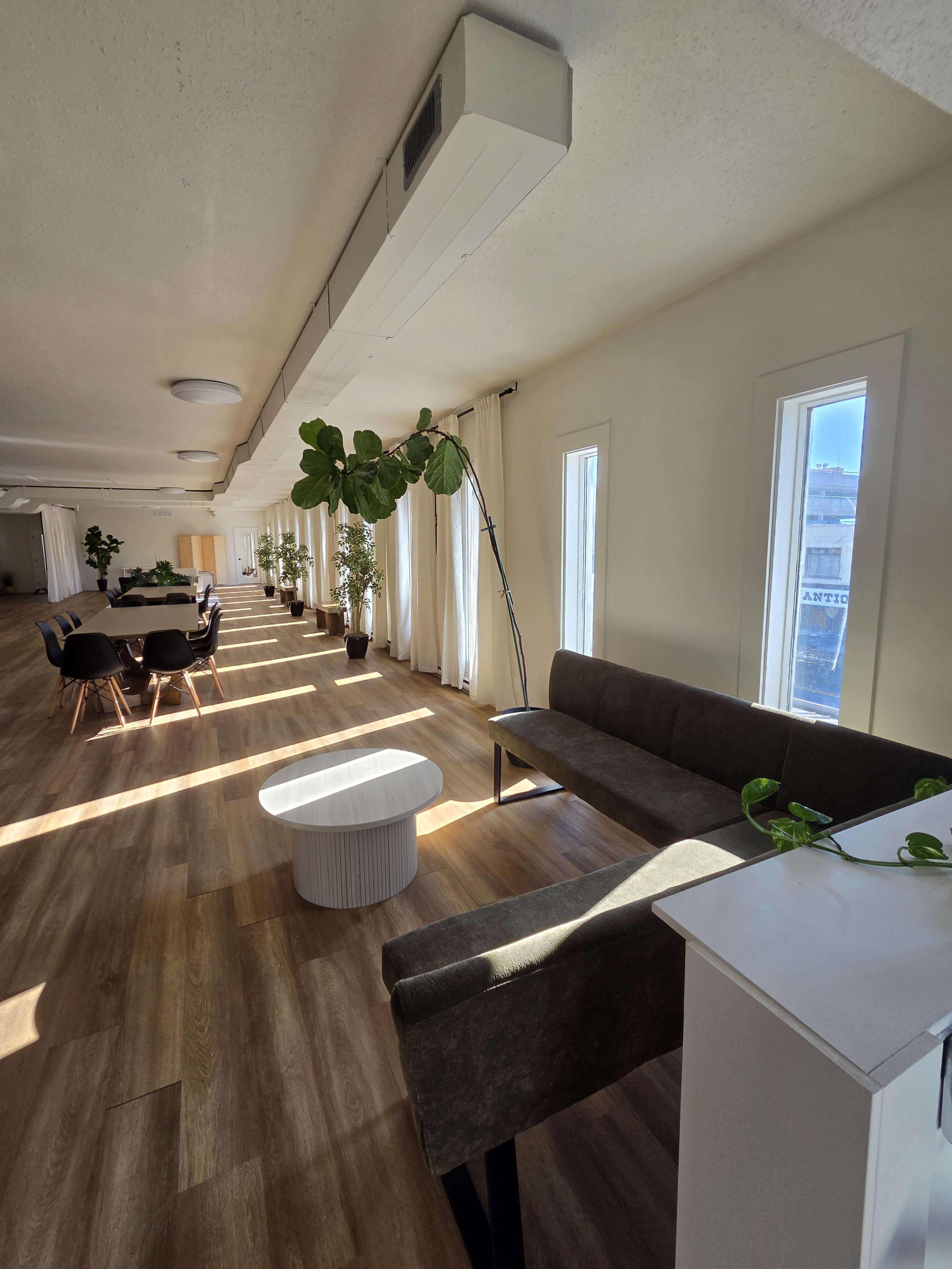 The image shows a modern, well-lit room with wooden flooring, featuring a long table set for dining, a few potted plants, and a seating area with a bench and a side table.