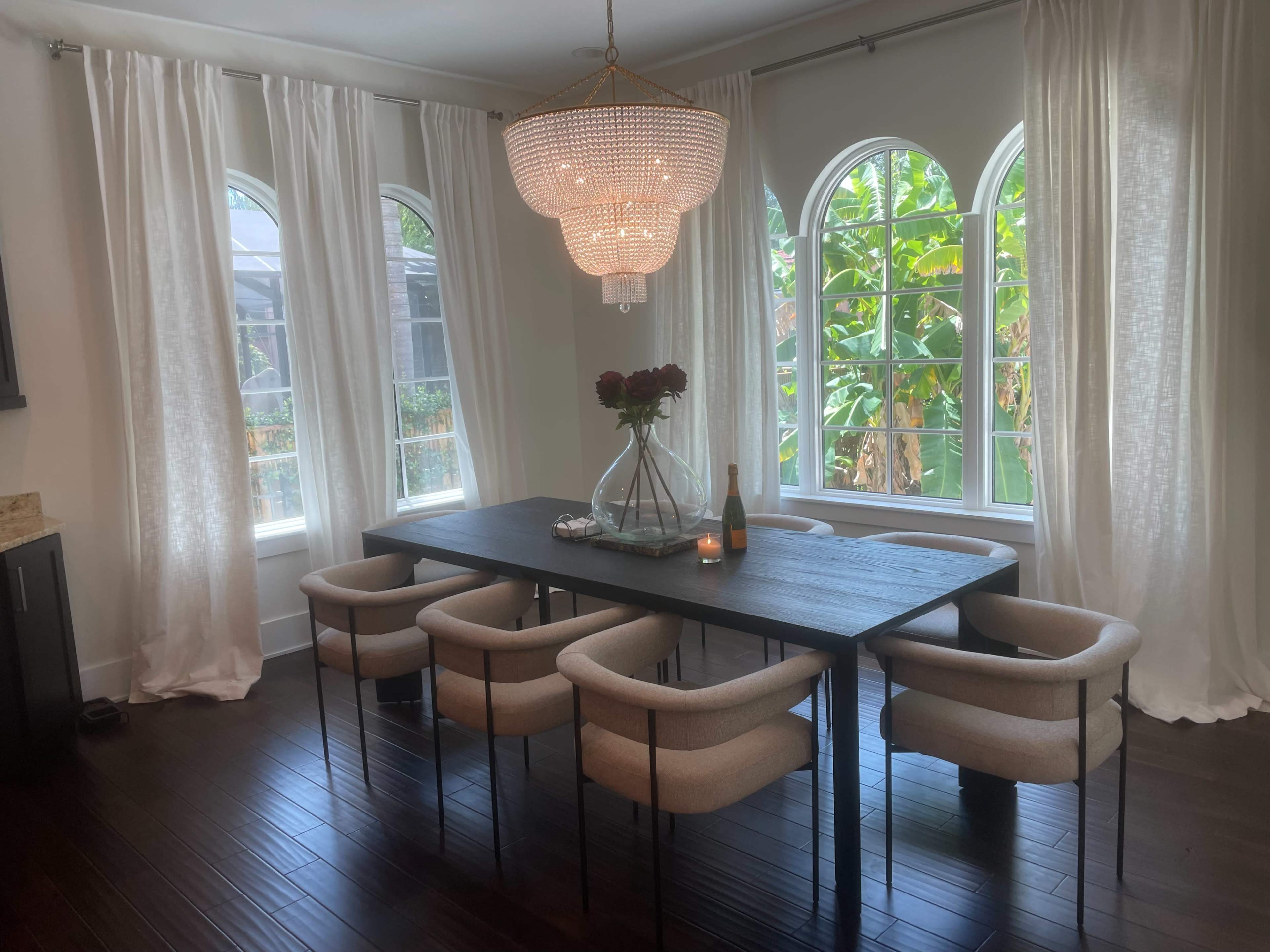 A dining area features a long wooden table surrounded by six upholstered chairs, with large arched windows and a chandelier overhead.
