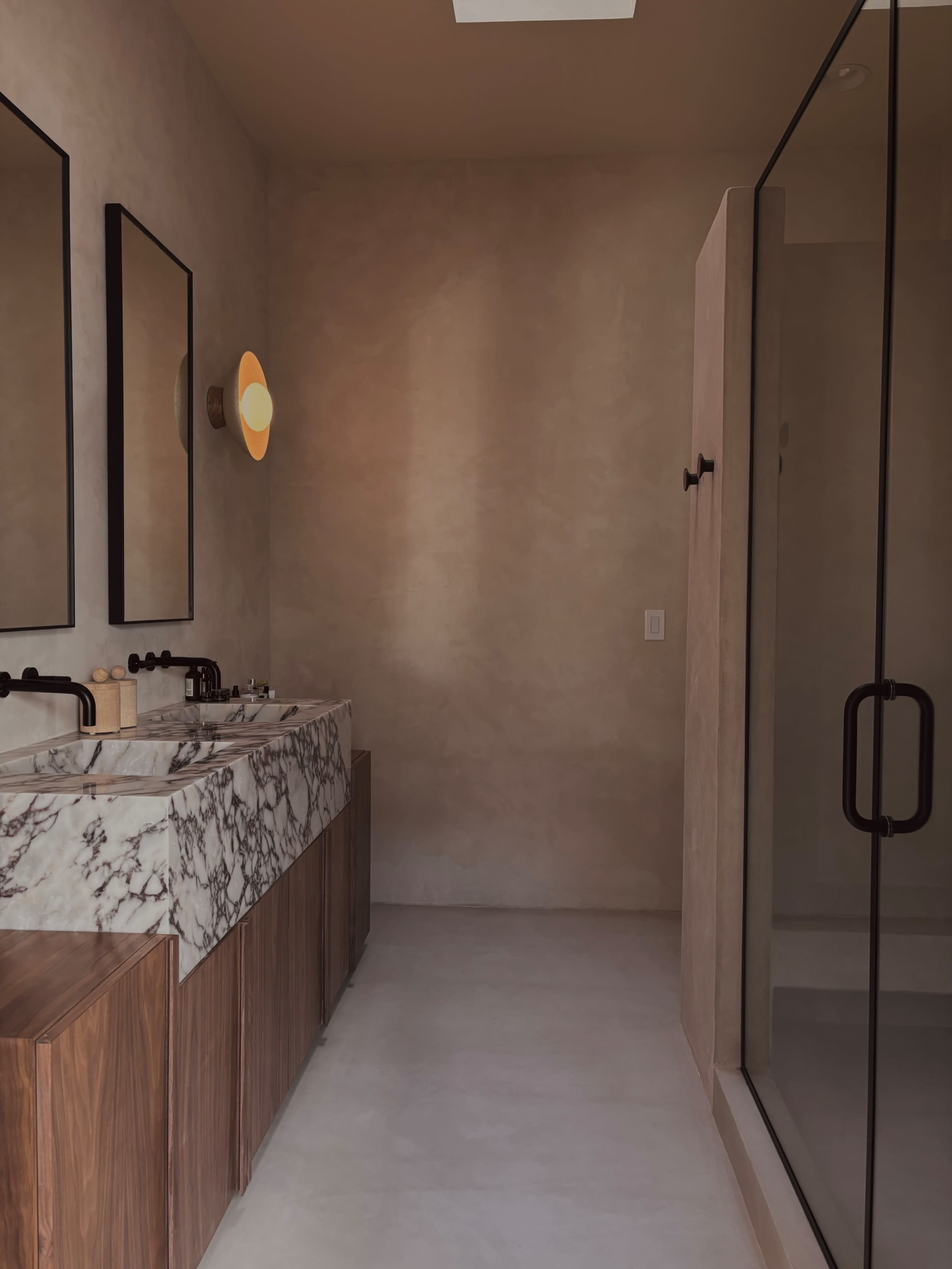 A modern bathroom with a marble double vanity, two mirrors, warm lighting, and a glass shower enclosure.