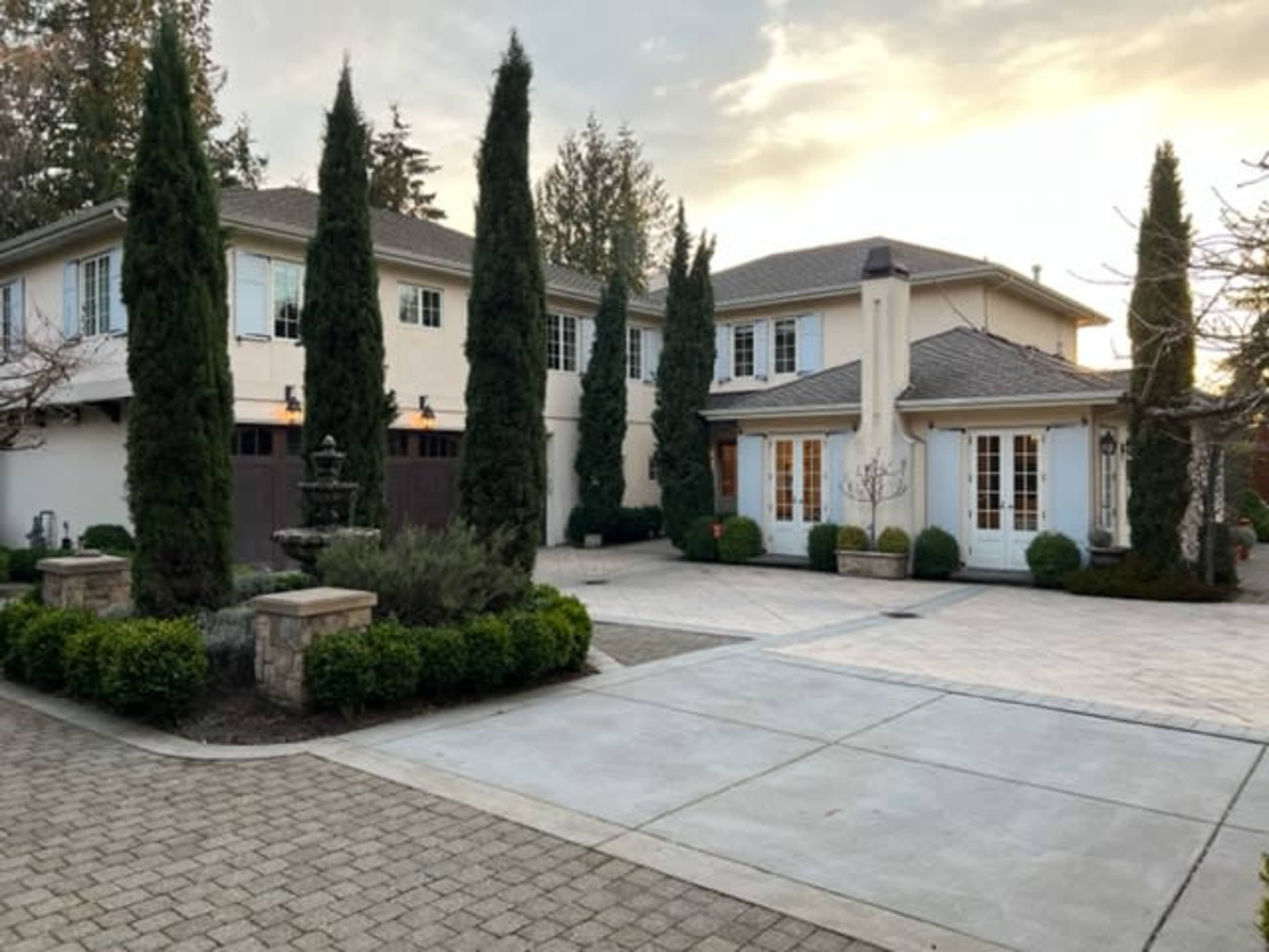The image shows a large, two-story house with manicured landscaping, featuring tall conifer trees flanking the entrance and a stone paved driveway leading to the garage.
