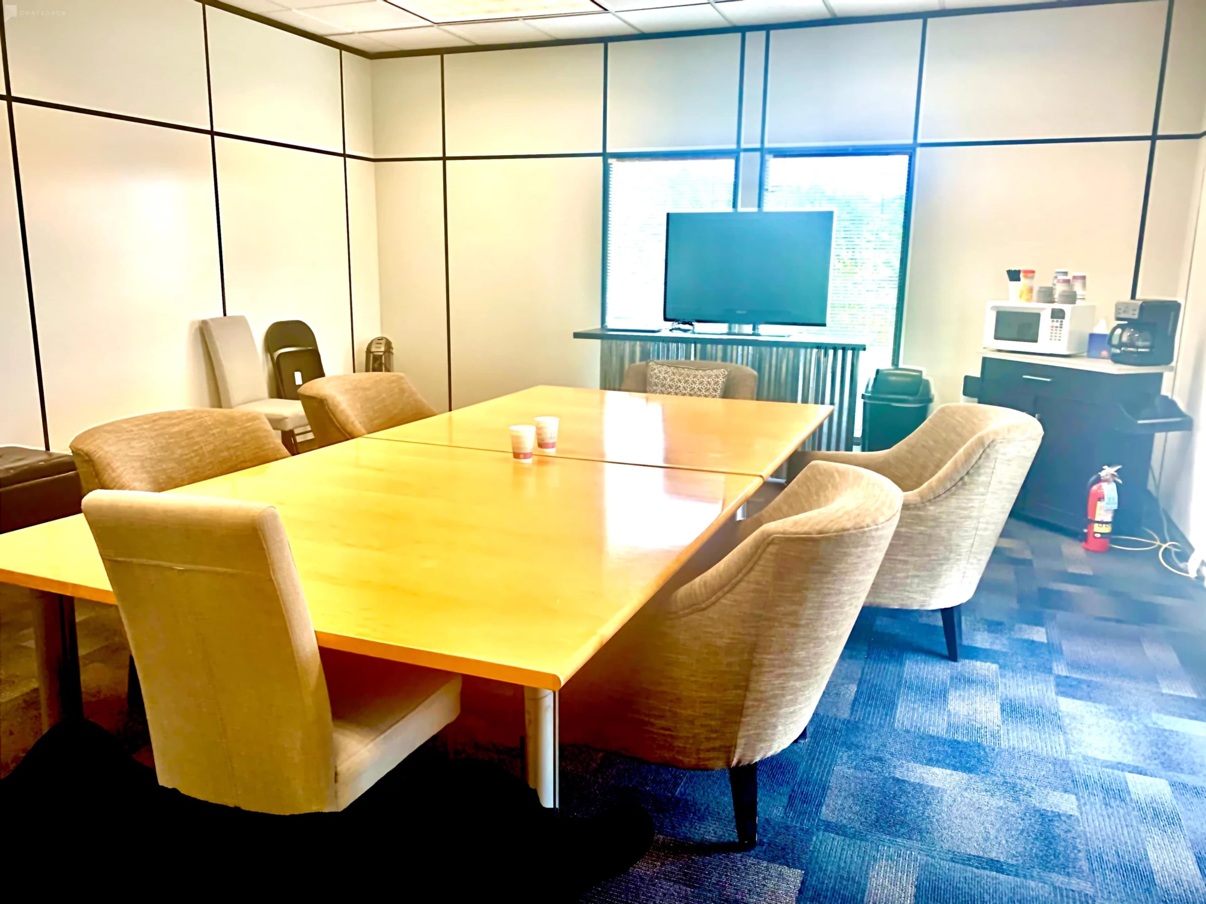 A conference room features a large wooden table surrounded by upholstered chairs, with a television and kitchenette visible in the background.