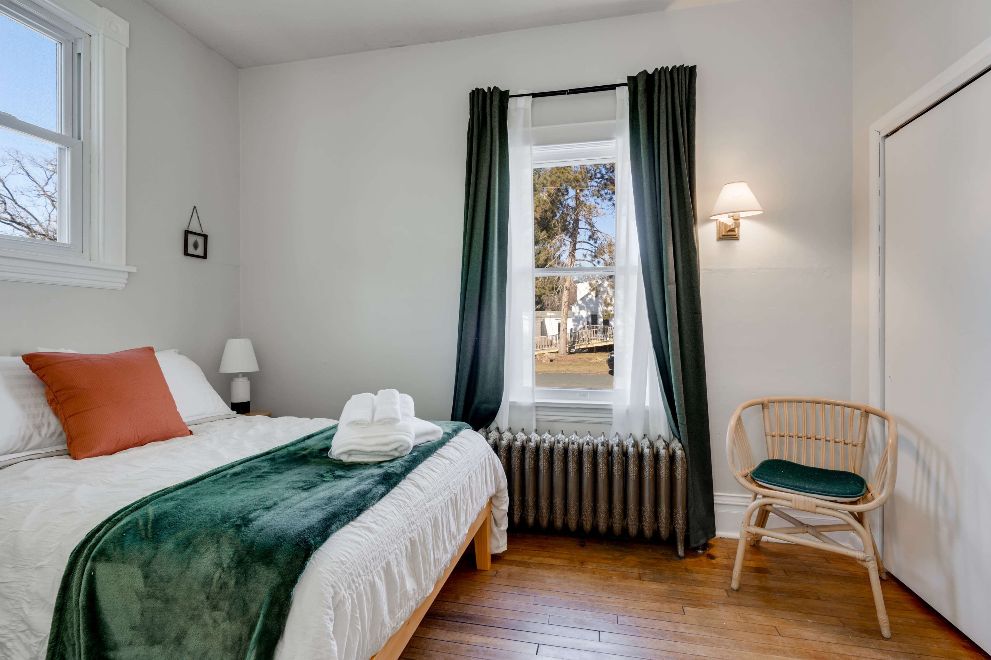The image shows a bedroom with a bed, a chair, and a window, featuring minimal decor and a radiator.