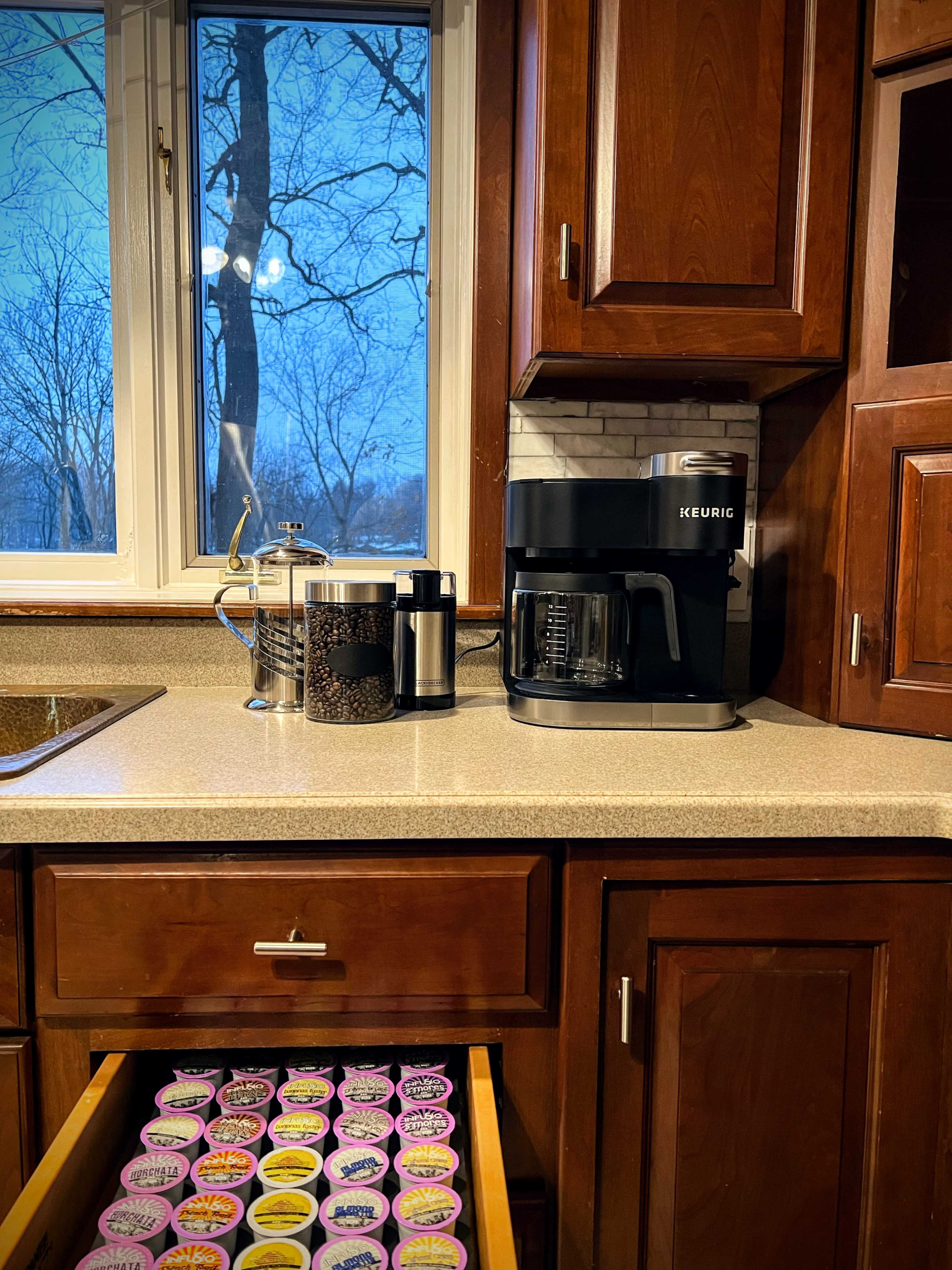 A kitchen counter displays a Keurig coffee maker beside a canister of coffee beans and a drawer filled with various coffee pods.