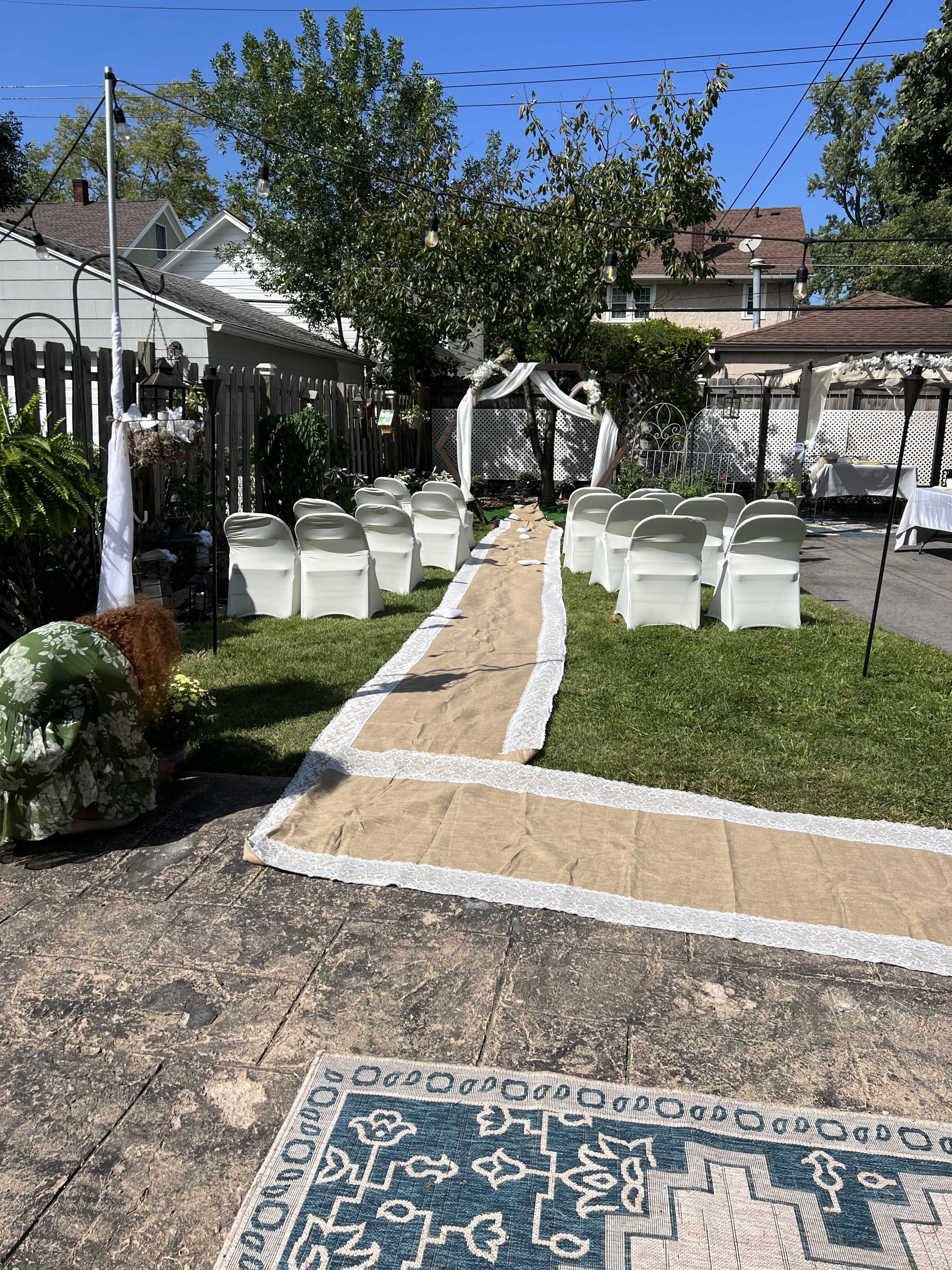 A wedding ceremony setup features white chairs arranged along a burlap pathway leading to a decorated arch under an outdoor tree.