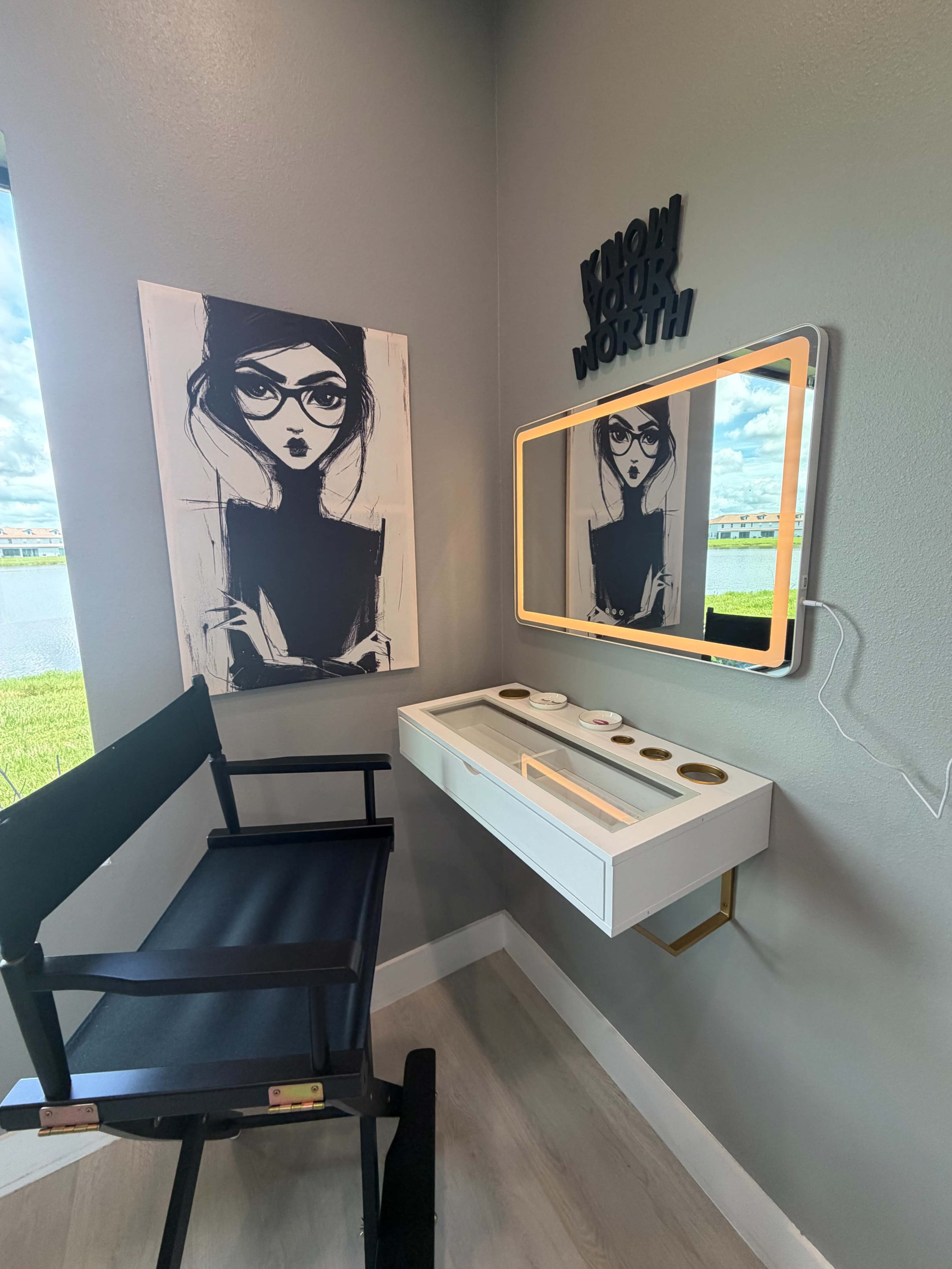 The image shows a makeup station with a chair, a wall-mounted mirror, and an artwork of a woman, located in a corner room with a view of a body of water.