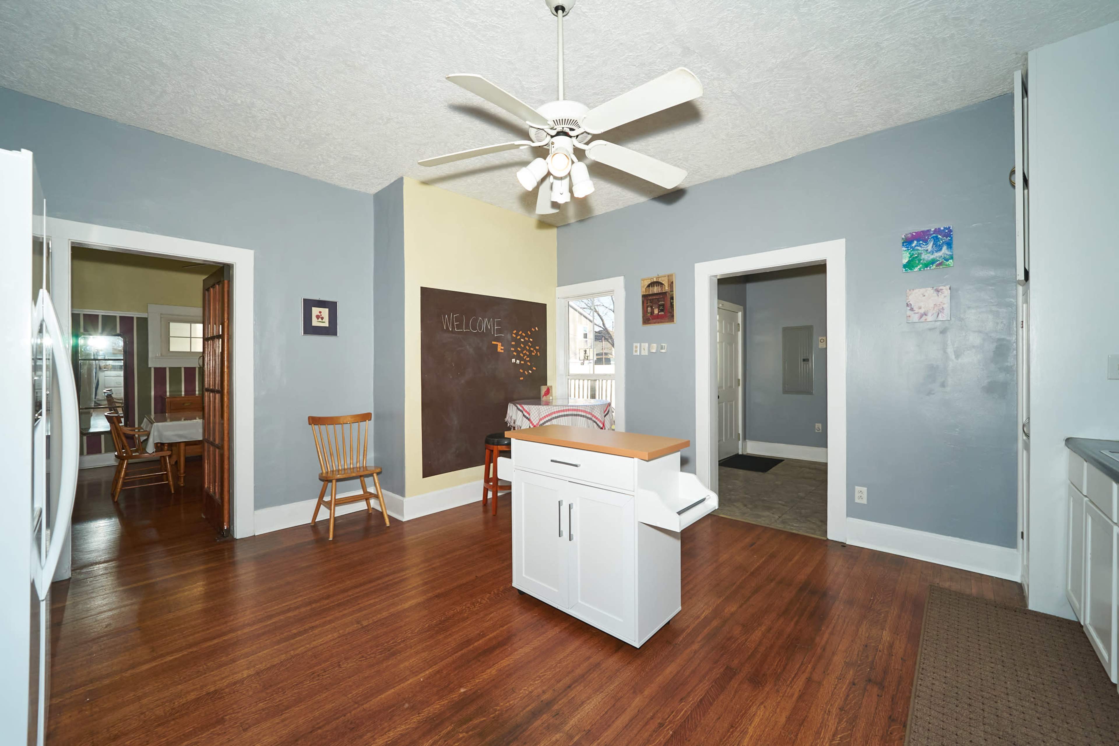 A spacious kitchen with a central island, a ceiling fan, and light blue walls, featuring a chalkboard on one wall and wooden floors.