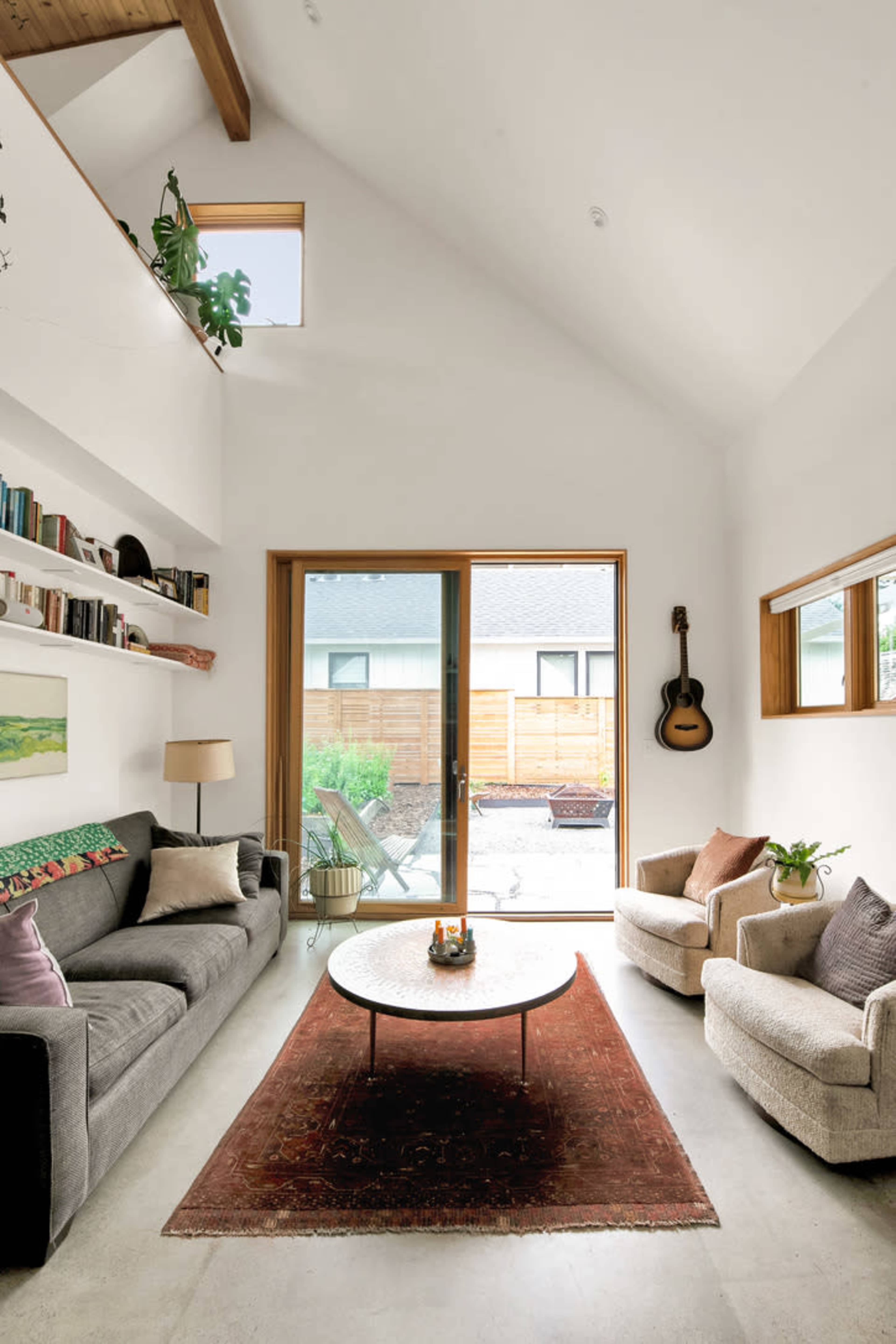A bright living room with a gray sofa, two light-colored armchairs, a round table, and large windows opening to a patio with outdoor seating.
