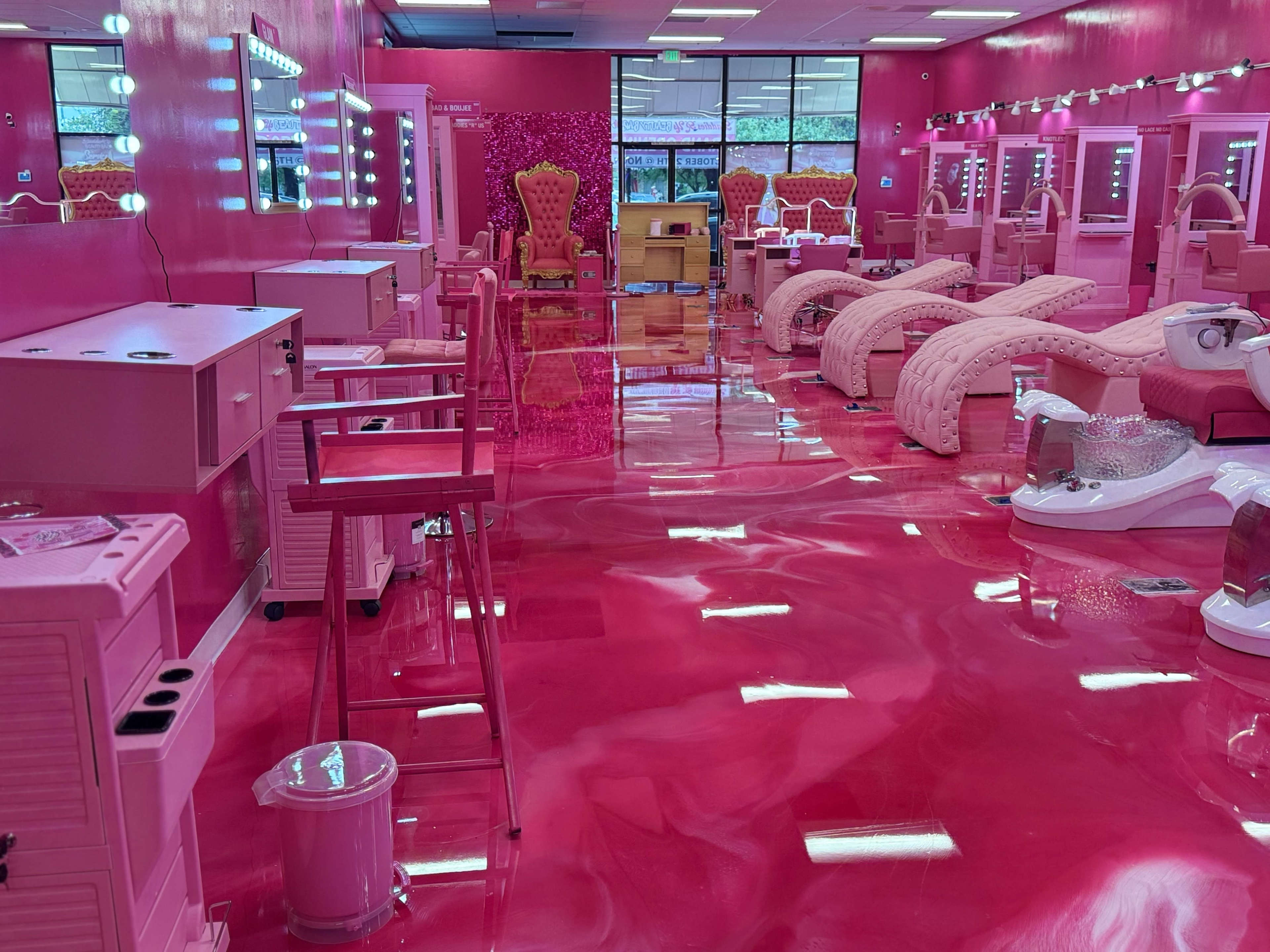 The image shows a nail salon interior featuring bright pink walls, furniture, and flooring, with multiple manicure stations and a reception area.