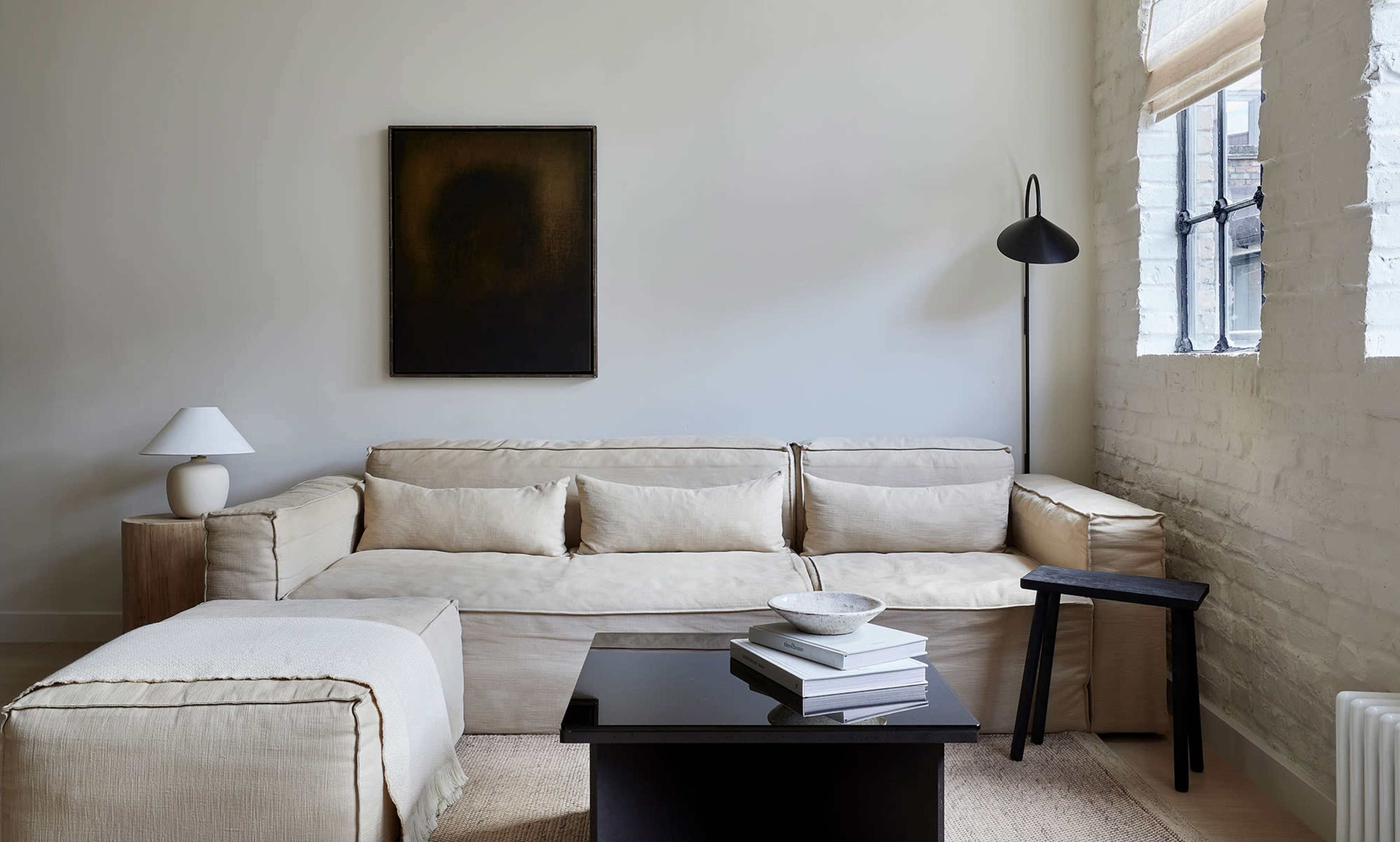 A minimalist living room with a beige sofa, a black coffee table stacked with books, and a lamp, against a backdrop of white walls and a single dark artwork.