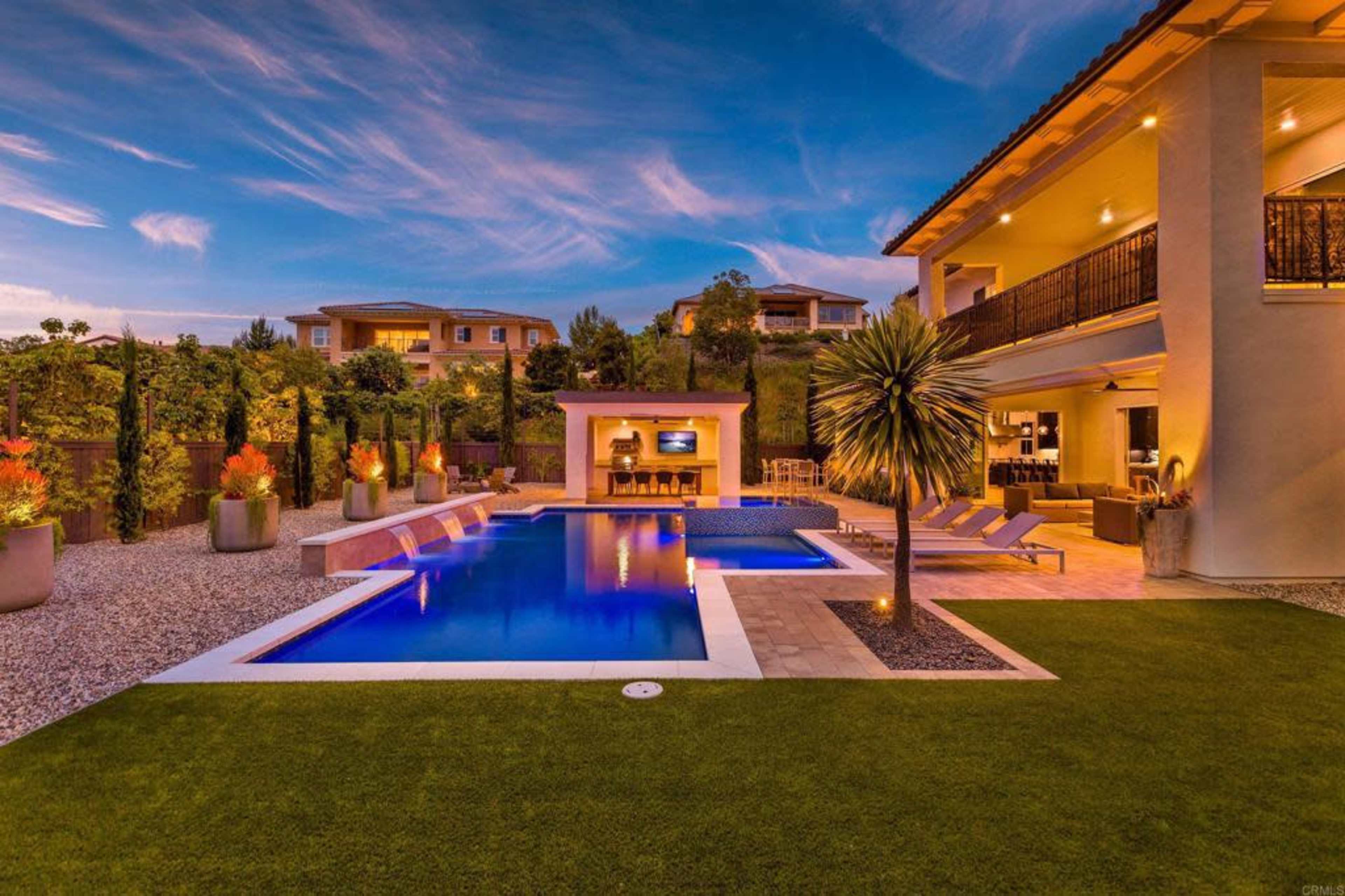 The image shows a modern backyard with a rectangular swimming pool, surrounded by lounge chairs and landscaped gardens, featuring a stone patio and an outdoor kitchen area.
