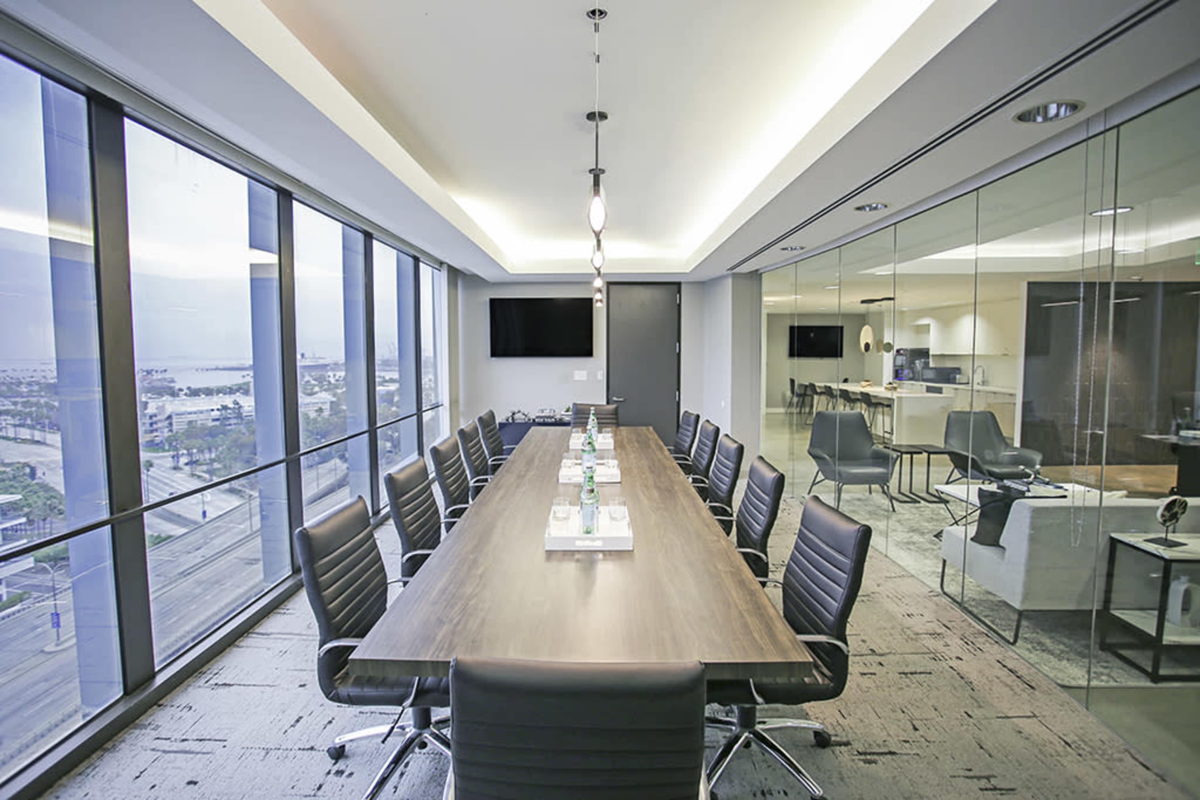 A long conference table with black chairs is set up in a modern meeting room featuring large windows overlooking a cityscape.