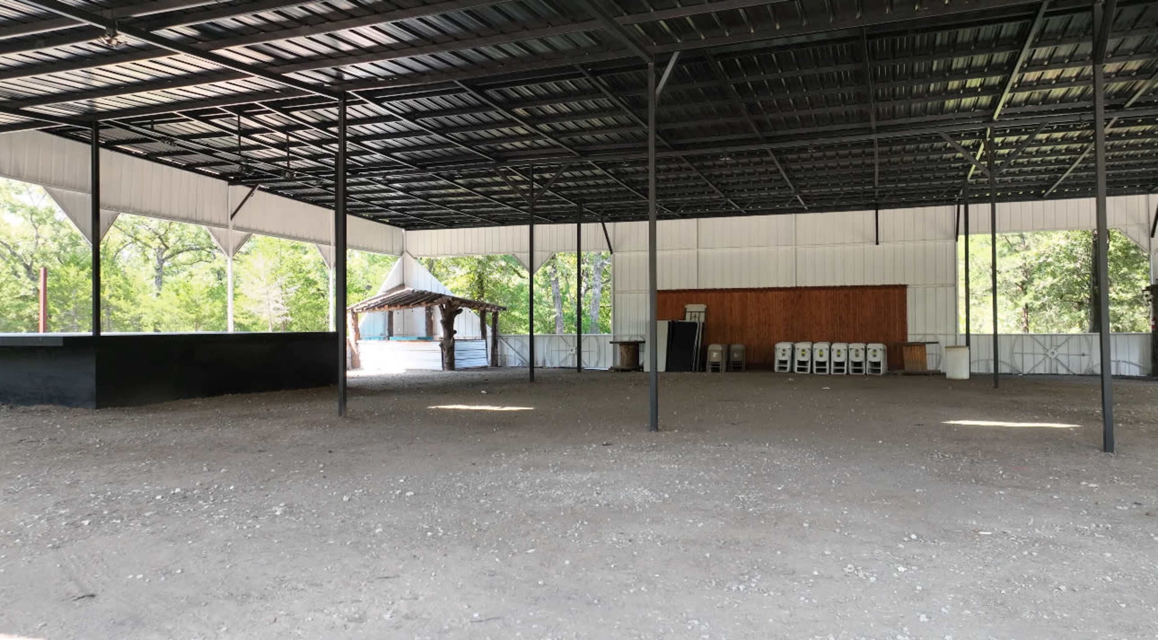 The image shows a large, empty indoor space with a metal roof, featuring open areas and a small structure resembling a gazebo in the background.
