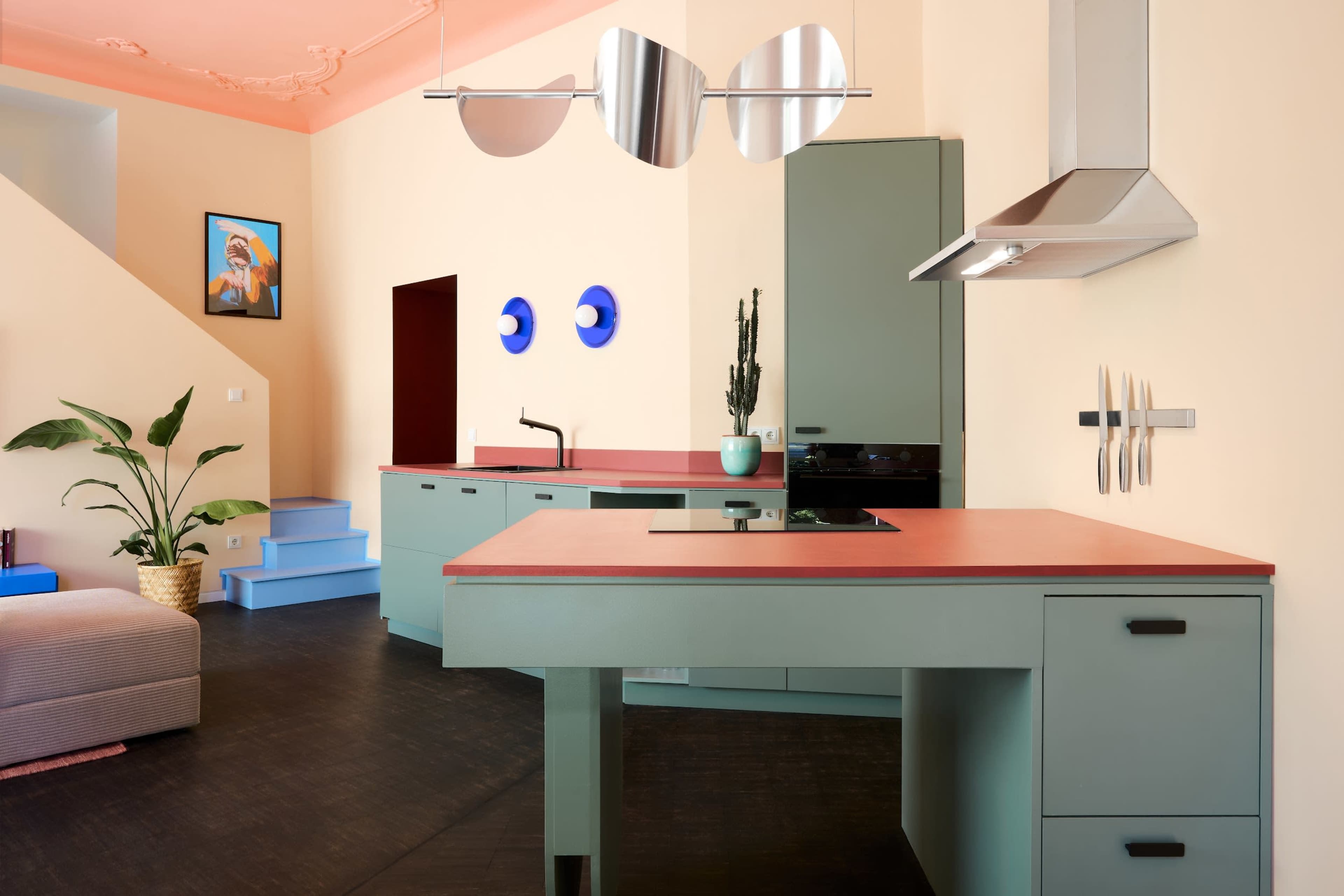The image features a modern kitchen with pastel green cabinets, a pink countertop, and a minimalist design, complemented by colorful wall art and a staircase.