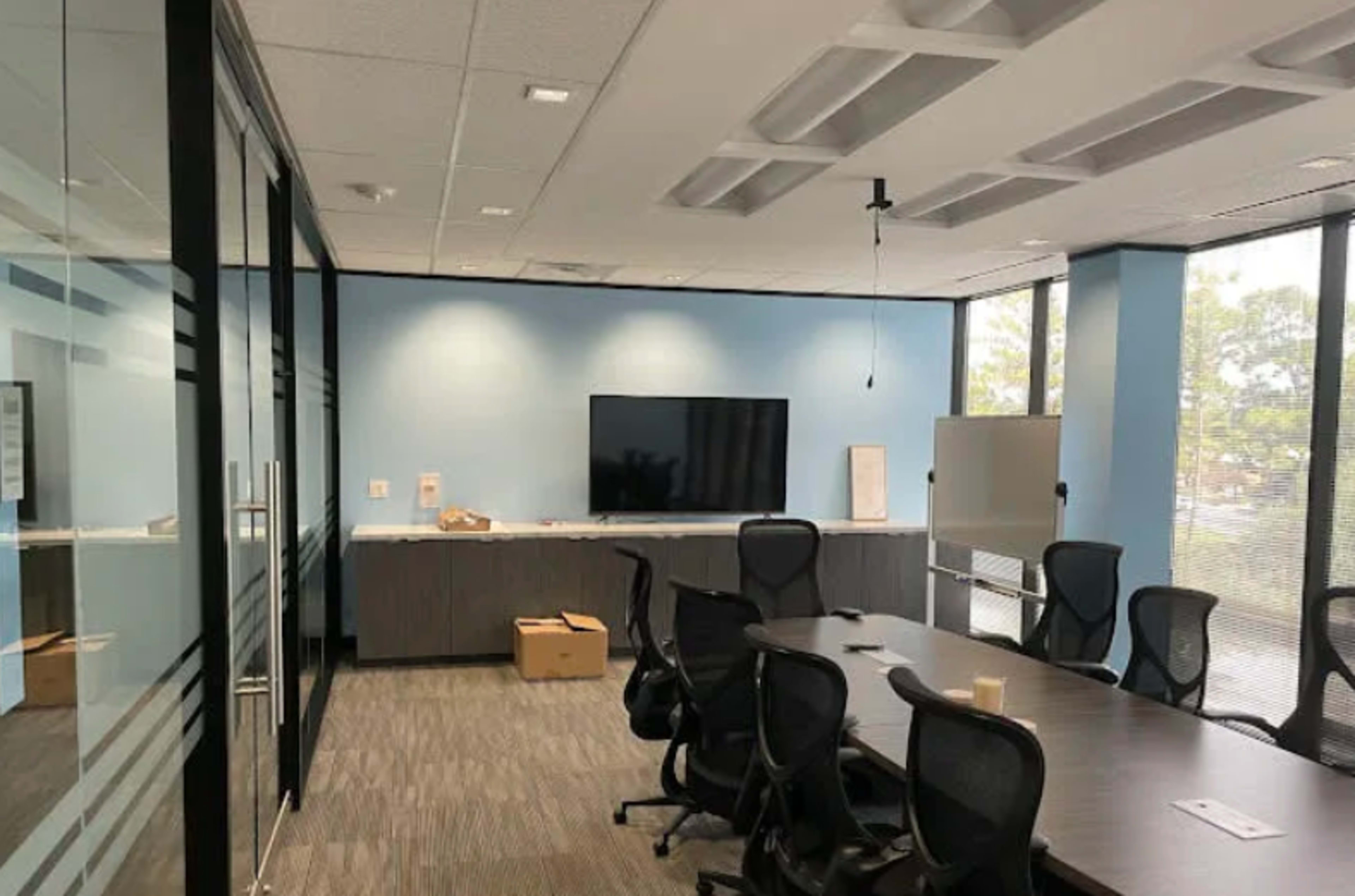 The image shows a modern conference room with a large table, ergonomic chairs, a wall-mounted TV, and a whiteboard, all set against light blue walls and large windows.