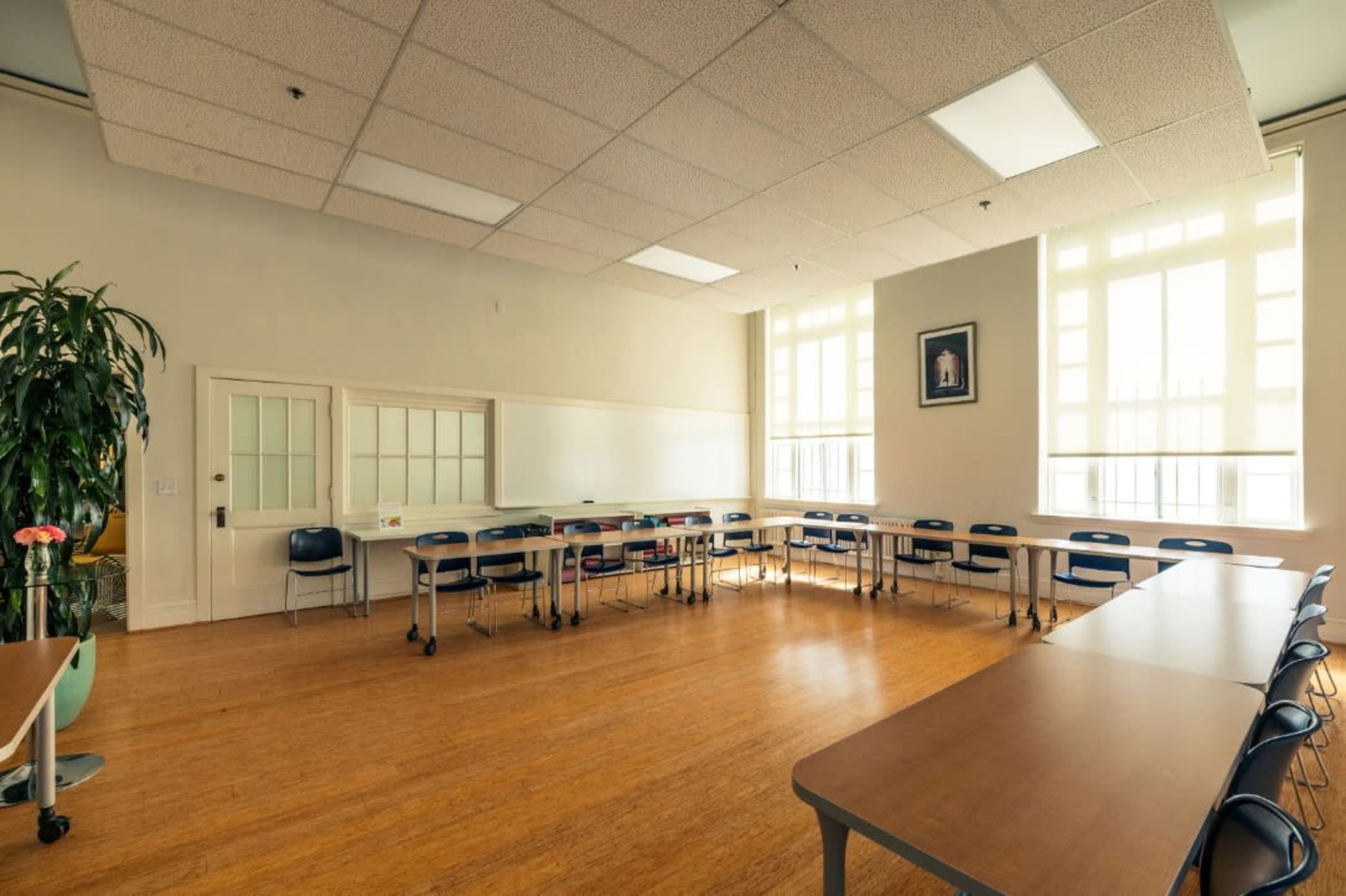 A bright classroom with wooden flooring, several tables arranged in a U-shape, and large windows allowing natural light to enter.