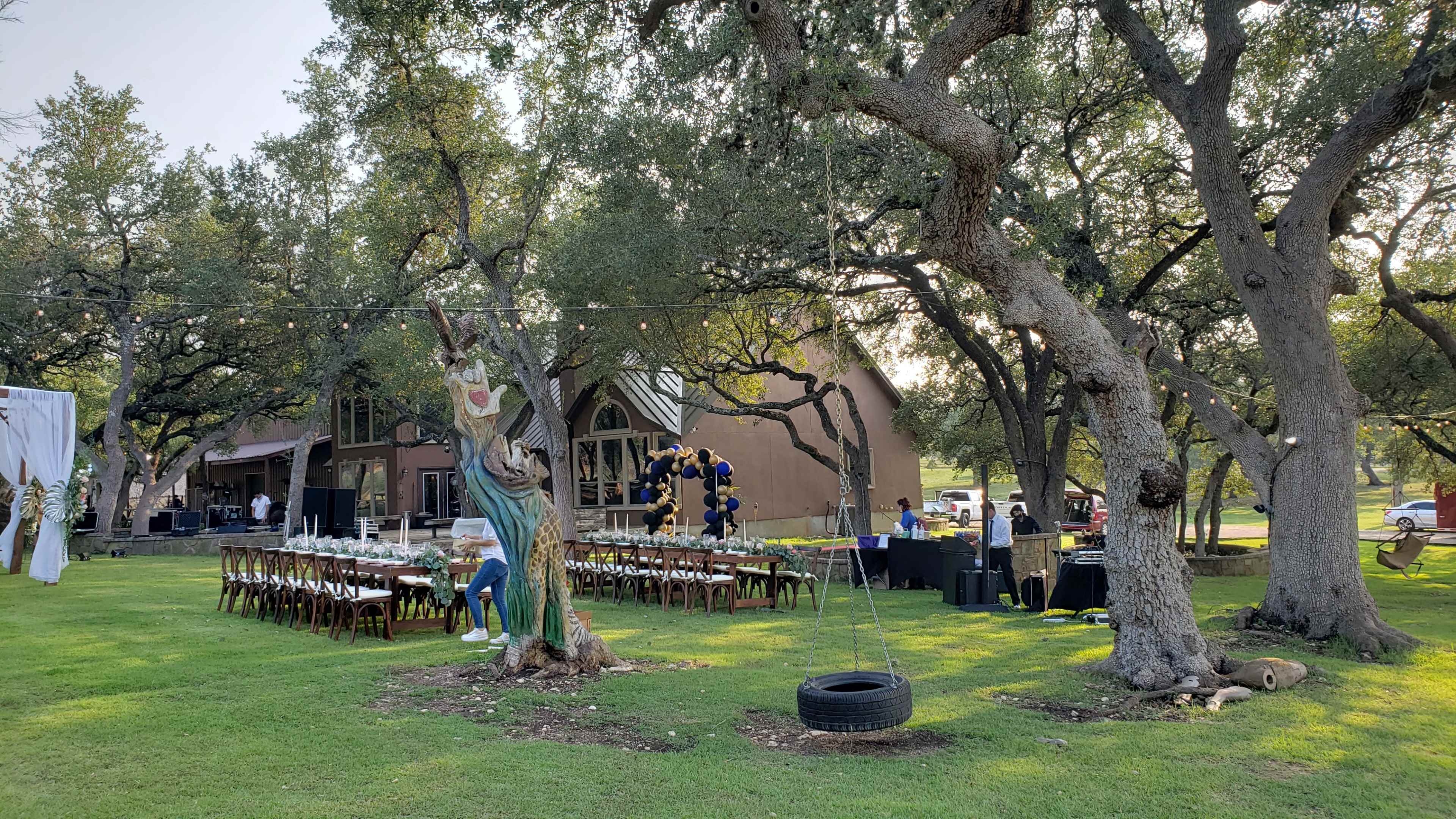A large outdoor event setup features a long table with chairs beneath trees, adjacent to a building and decorated areas with balloons.