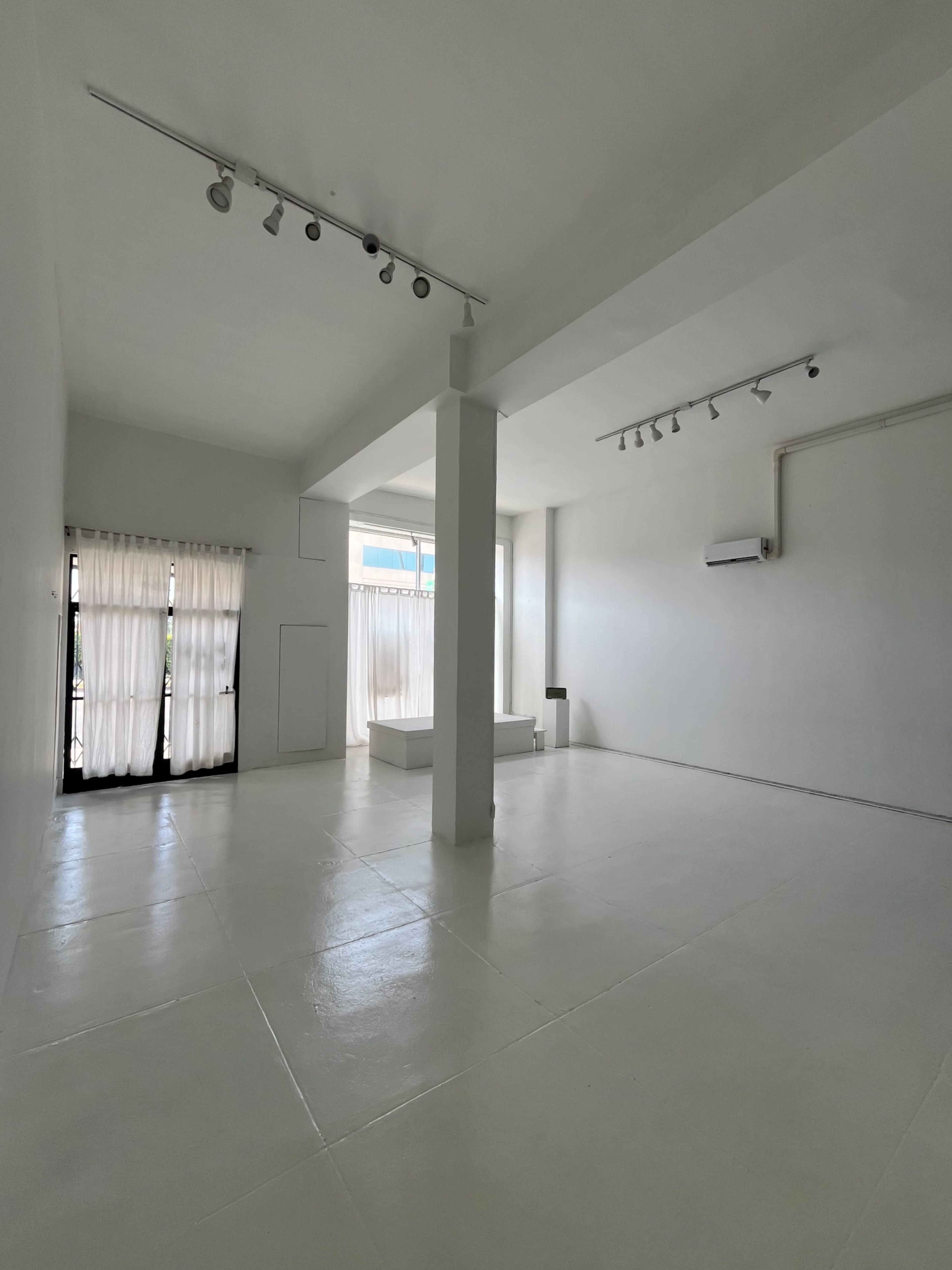 The image shows a spacious, empty room with white walls and floor, two large windows covered by sheer curtains, and track lighting on the ceiling.