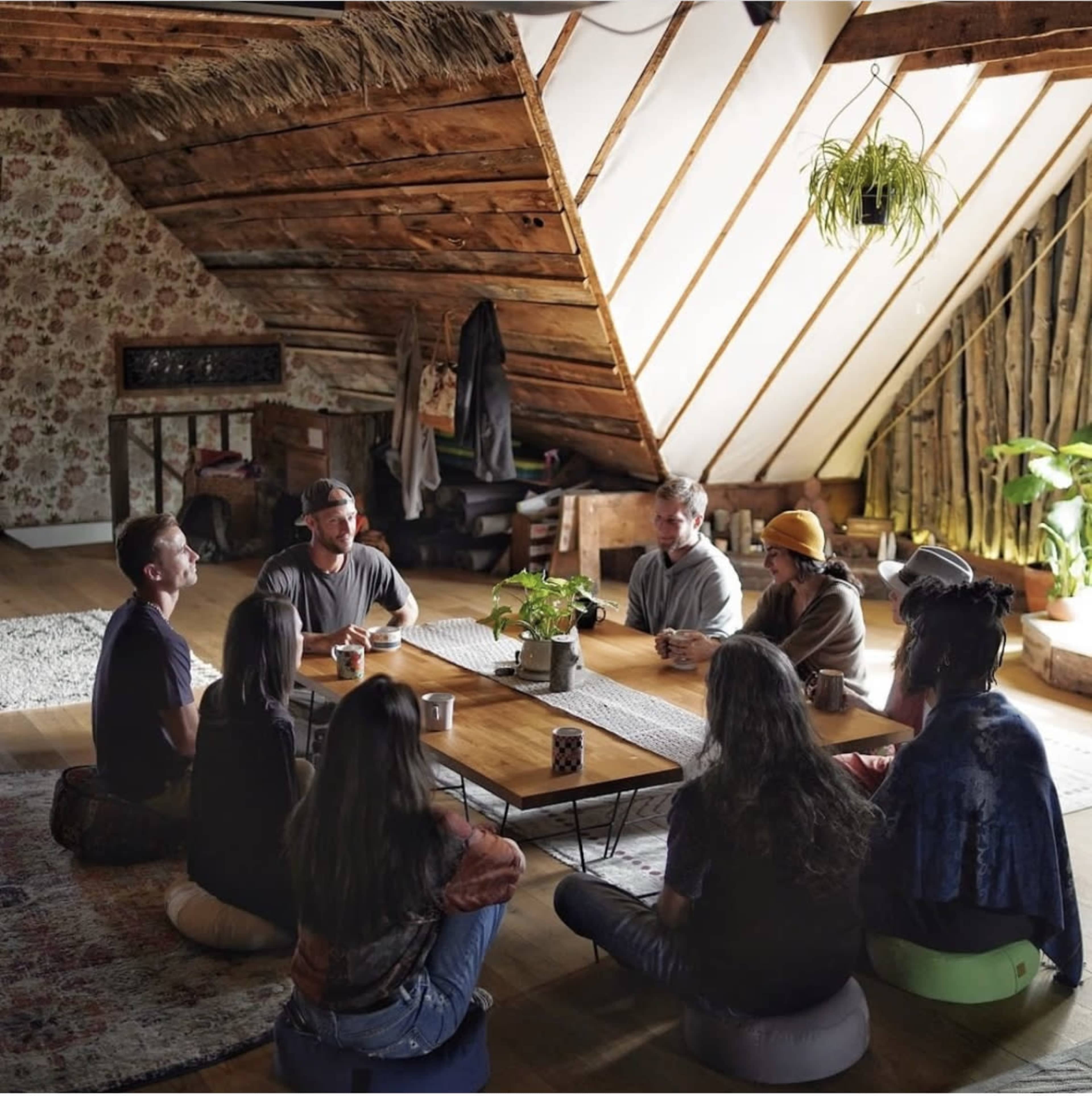 A group of people gathers in a warmly lit attic space, seated on cushions around a wooden table surrounded by plants and rustic decor.
