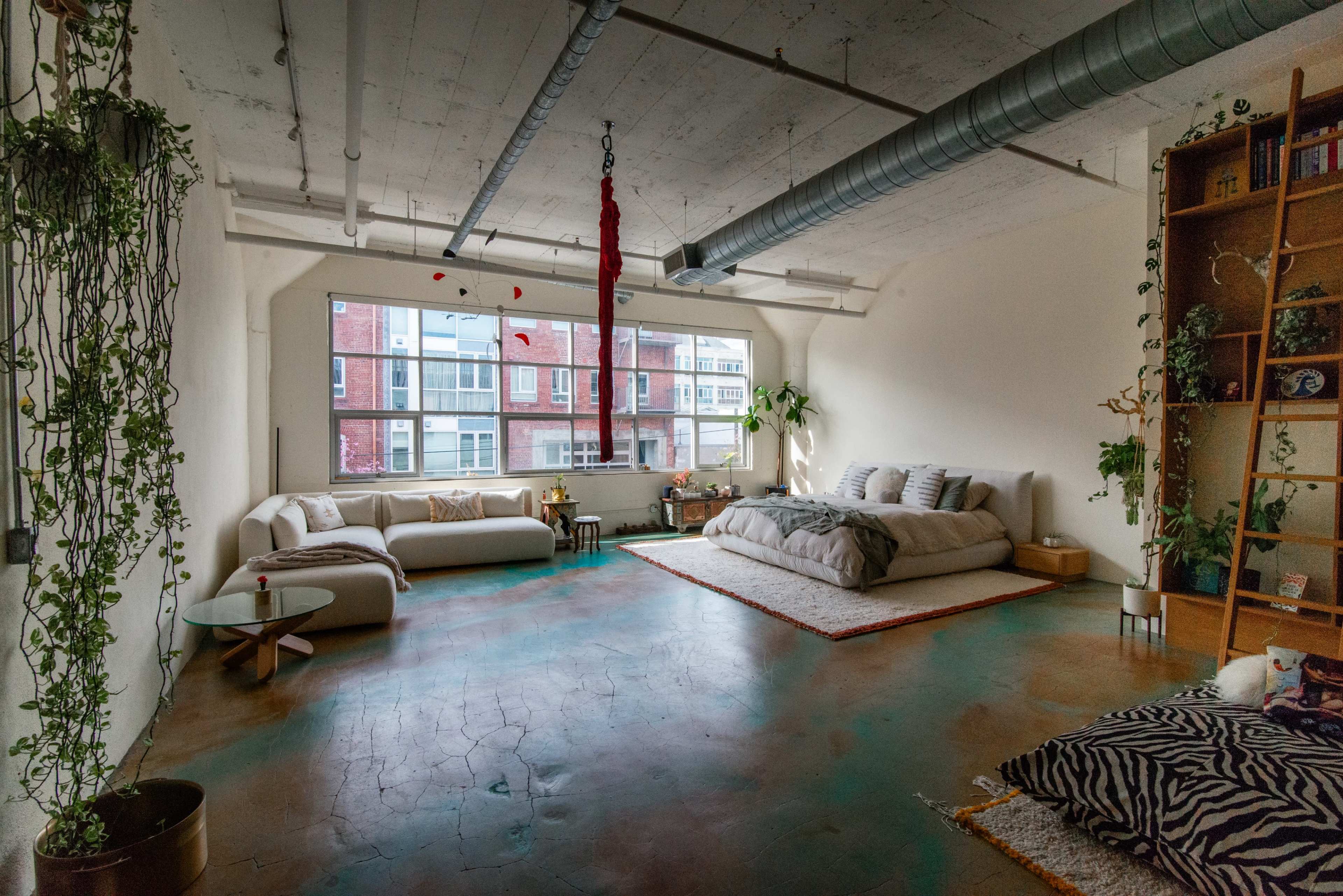 The image shows a spacious, minimalist living area with a bed, a large sofa, and plants, arranged in front of a large window.