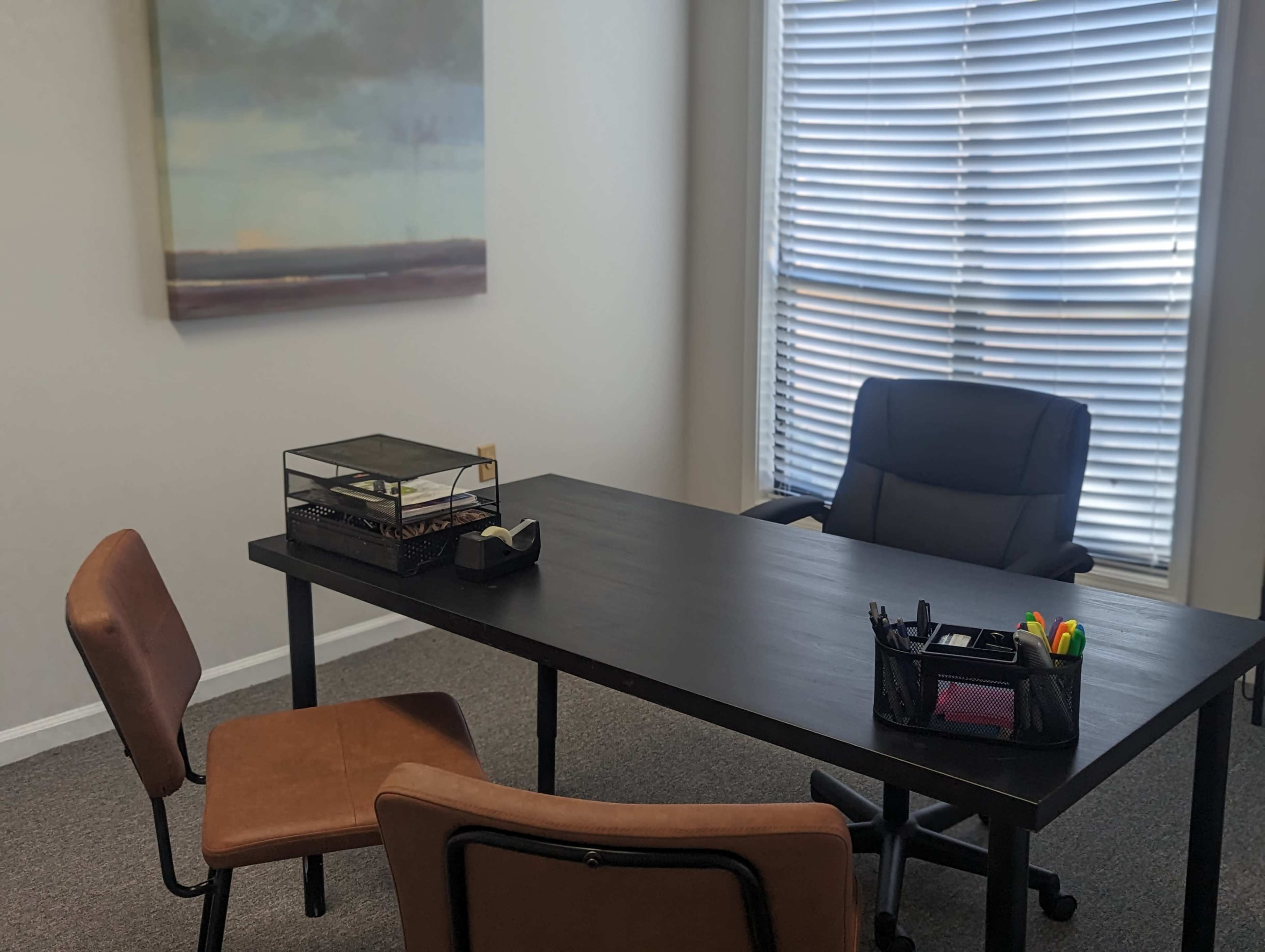 A clean office interior features a black desk with a basket of supplies and two brown chairs, alongside a window with vertical blinds and a painting on the wall.