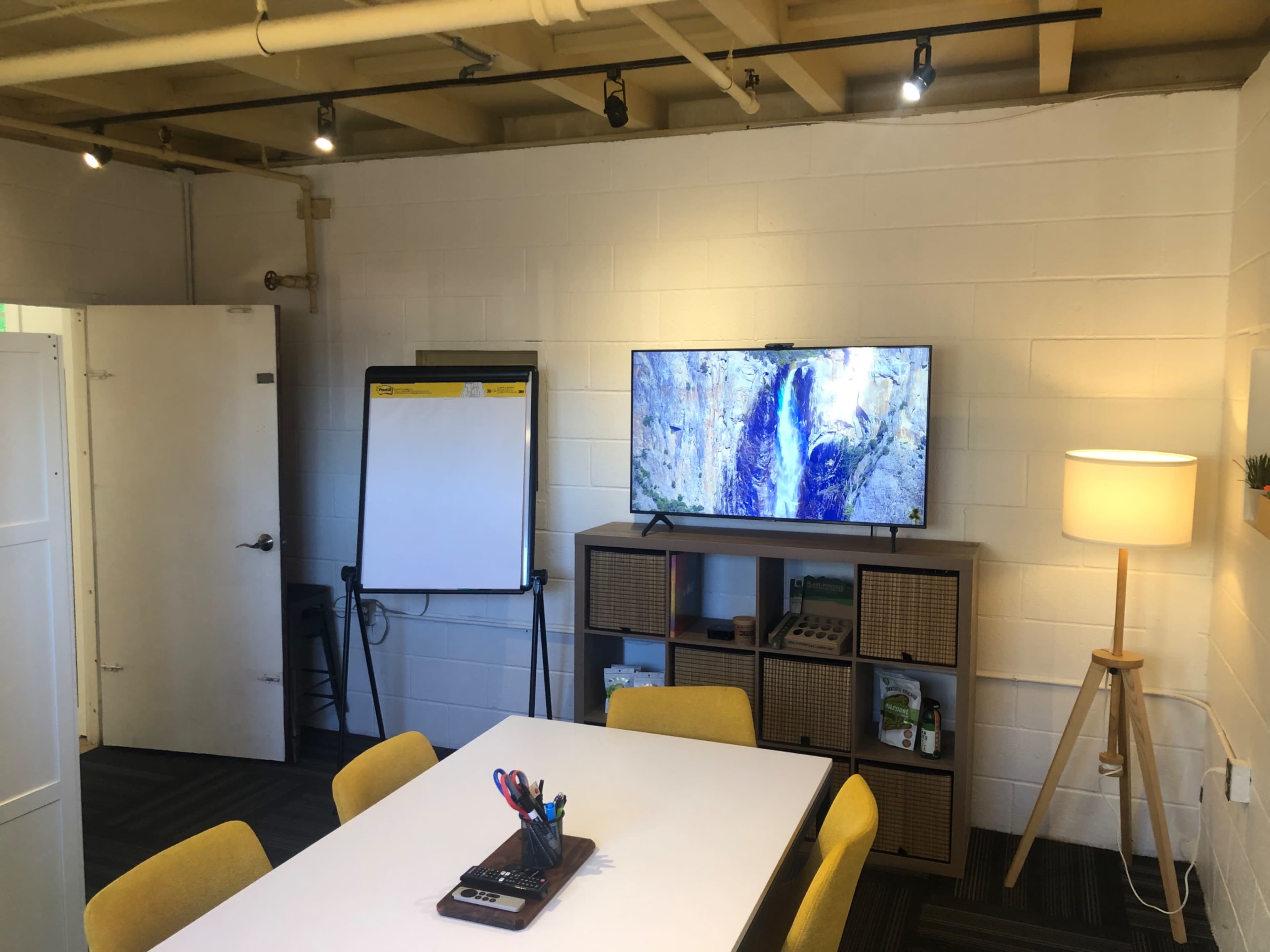 A small meeting room with a whiteboard, a television displaying a natural scene, a shelving unit, a table with pens and notebooks, and a floor lamp.