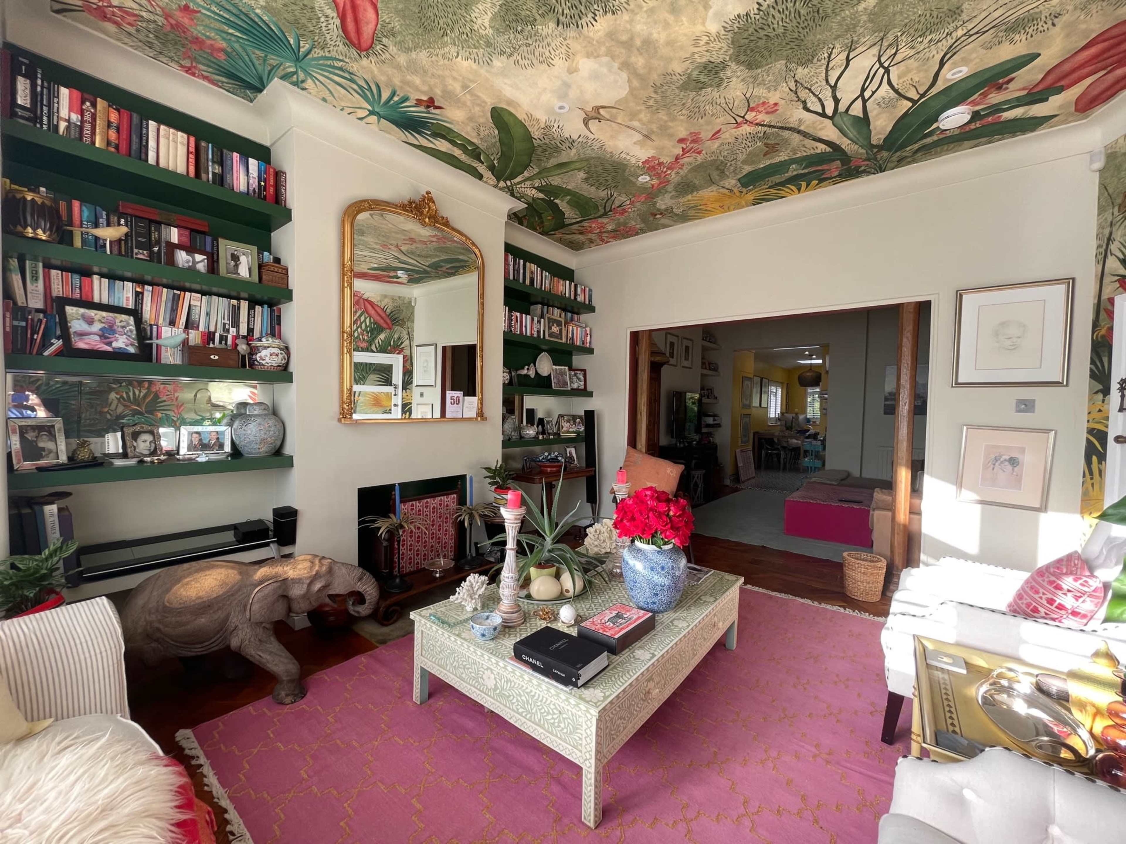 A vibrant living room features a floral ceiling mural, bookshelves filled with books, a mirrored wall, and a coffee table adorned with decorative items and flowers.