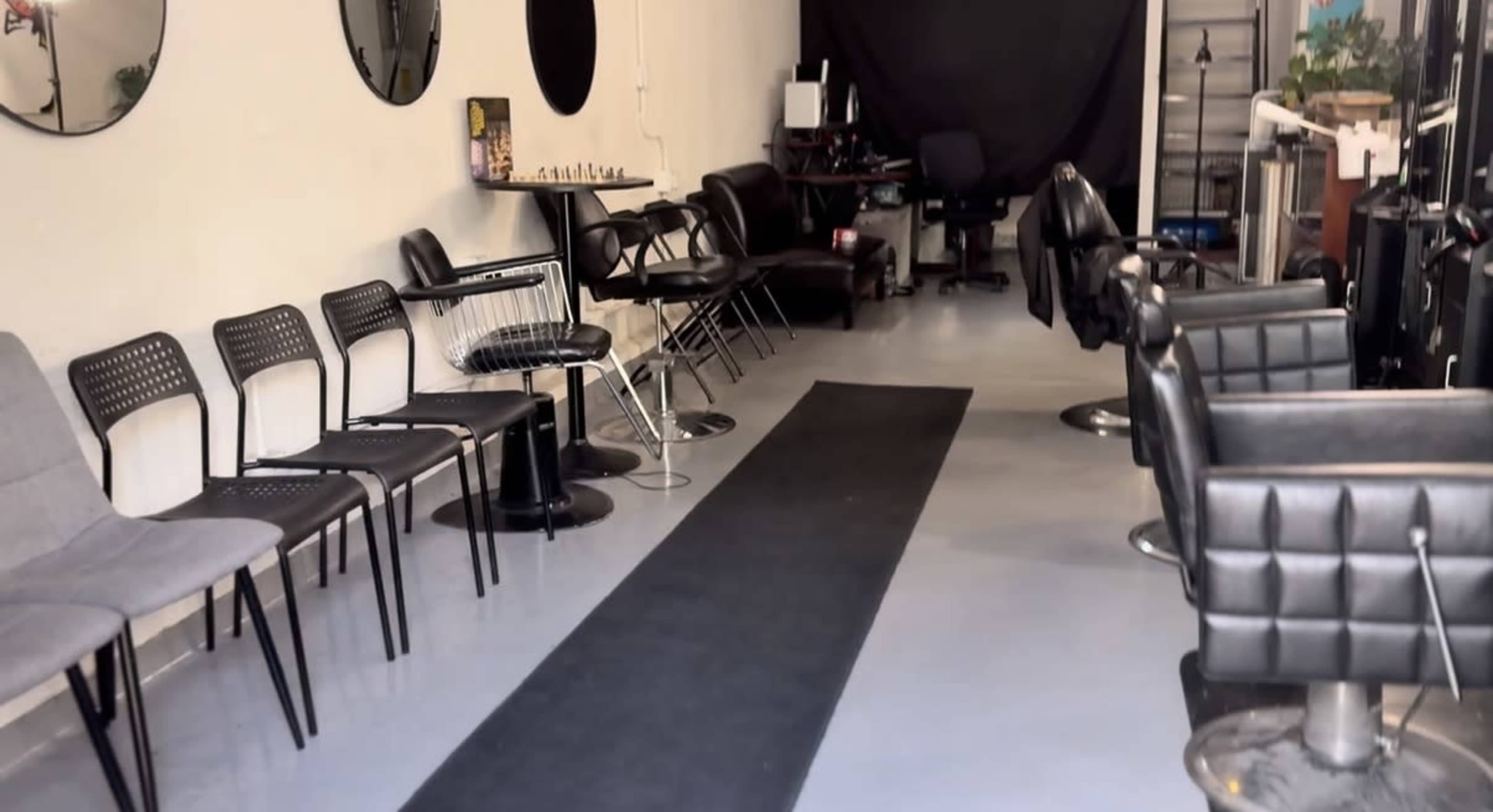 The image shows a barber shop interior with various black chairs, a long black mat, and mirrors on the walls.