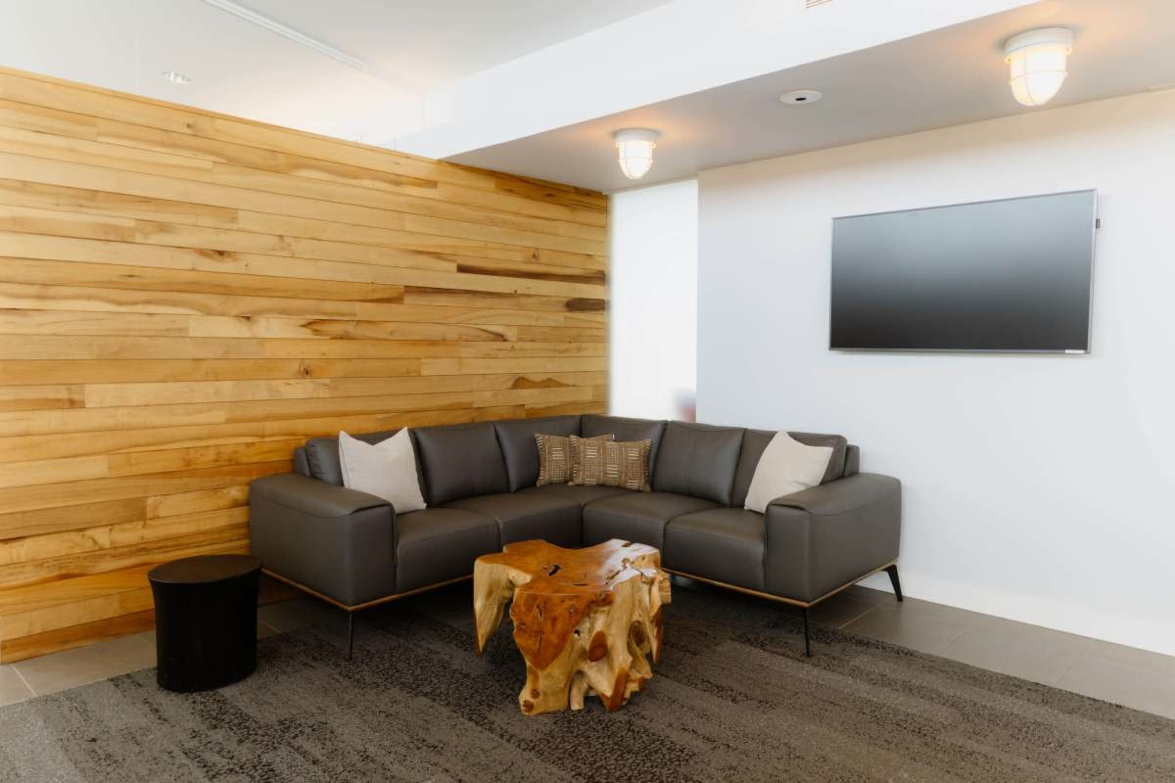 The image shows a modern seating area featuring a dark gray sectional sofa, a natural wood coffee table, and a wooden accent wall, with a flat-screen television mounted above.