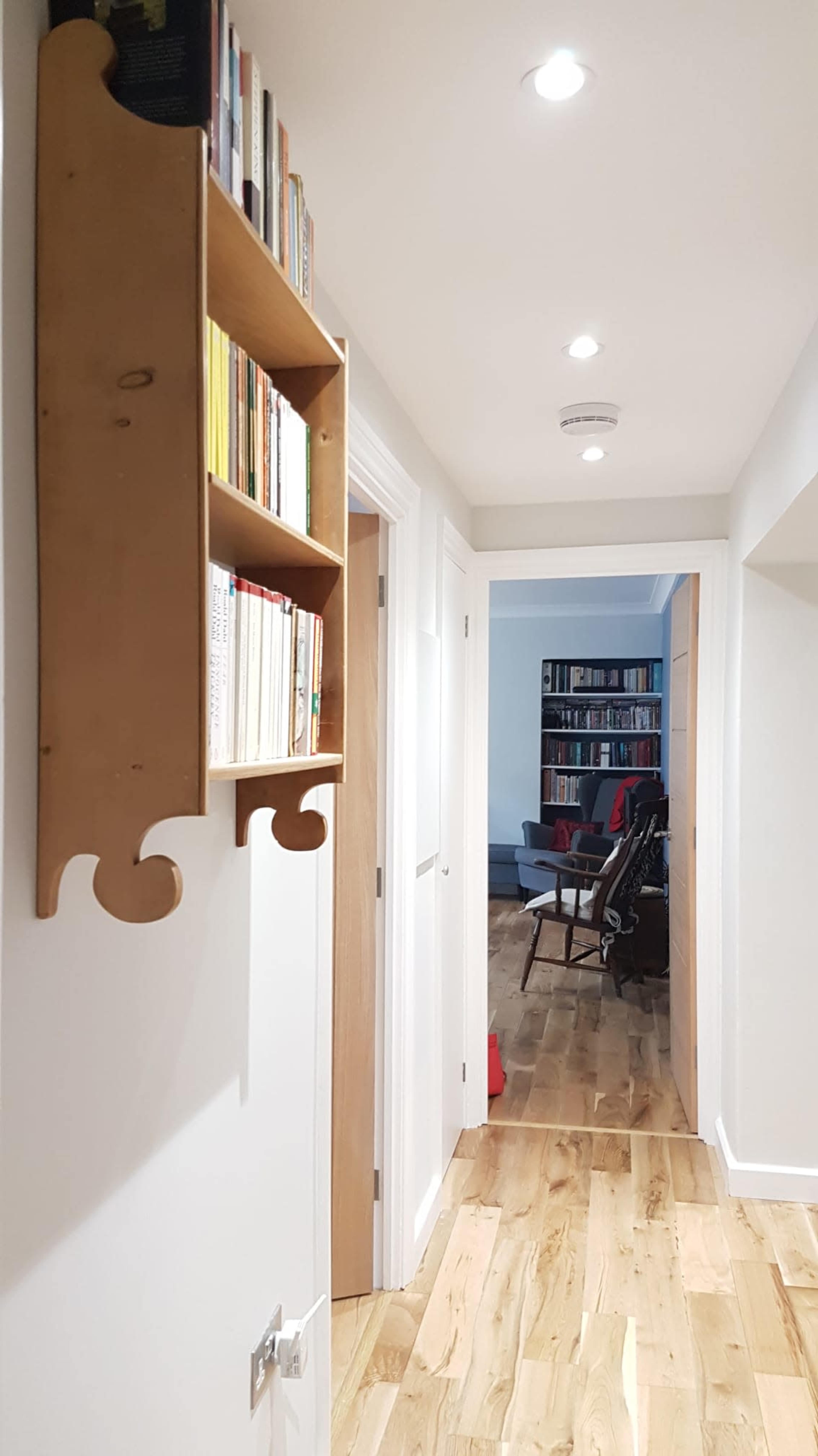 A narrow hallway features a wooden bookshelf mounted on the wall, leading to a room with additional shelves filled with books.