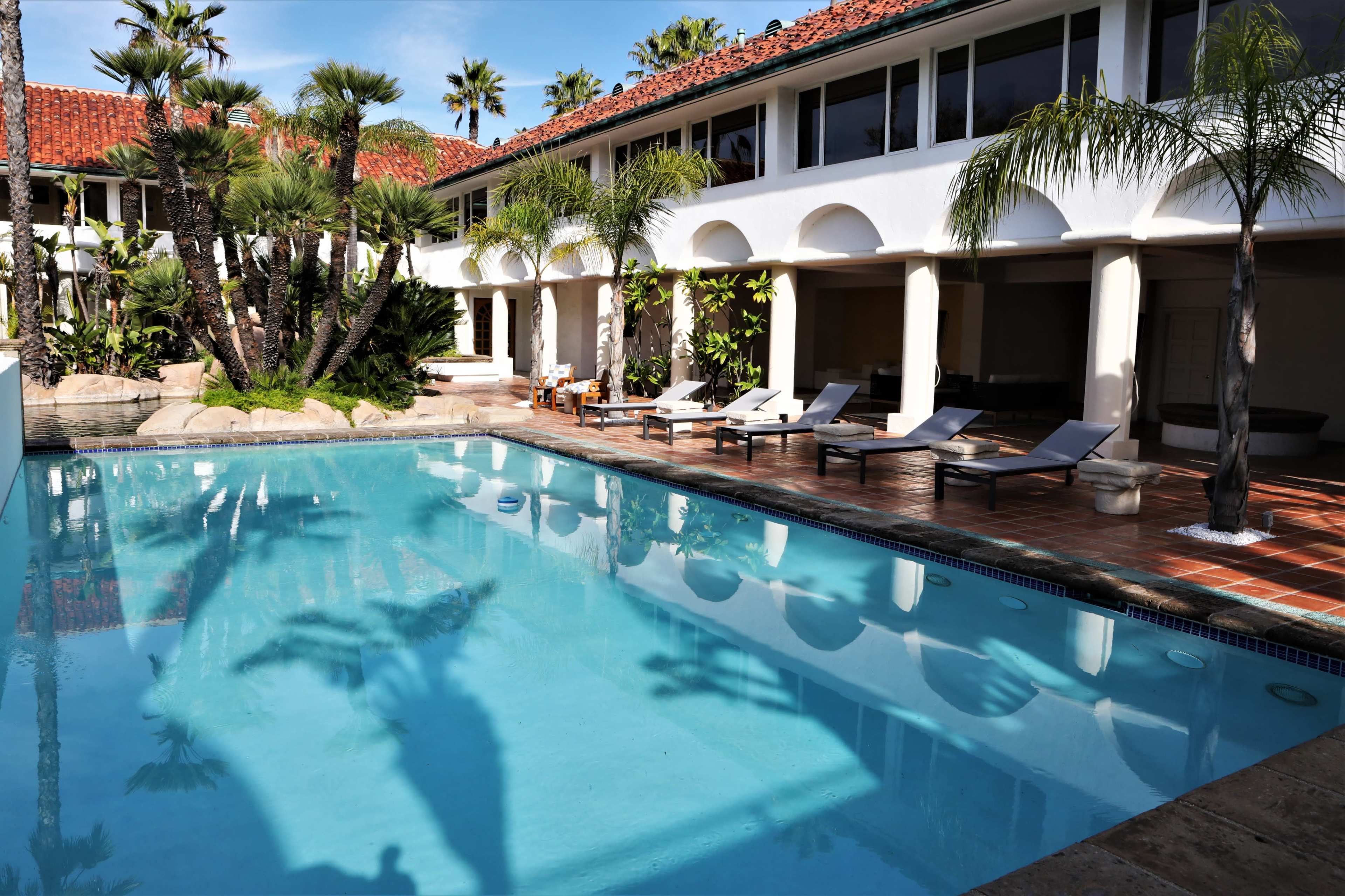 A swimming pool surrounded by lounge chairs and palm trees, with a two-story building in the background.